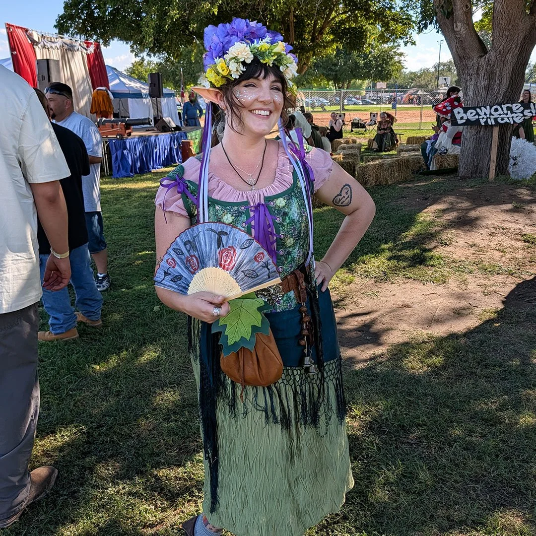 Emily, soprano vocalist for Irish folk duo Secret Moss Meadow, smiling in a floral crown and Renaissance festival attire holding a hand fan.