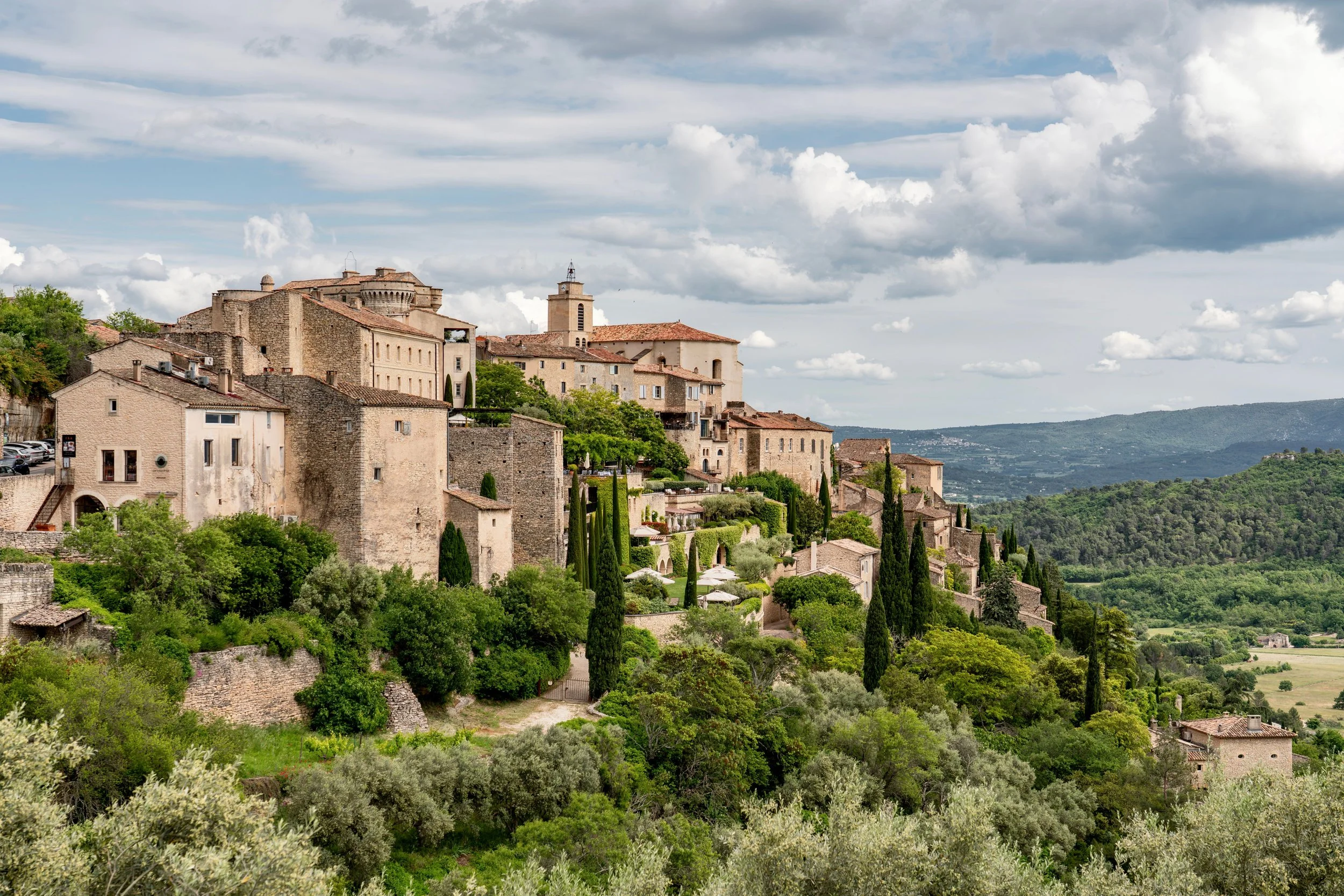 A scenic view of an Italian hillside town with stone buildings, red-tiled roofs, green trees, and rolling hills under a partly cloudy sky.