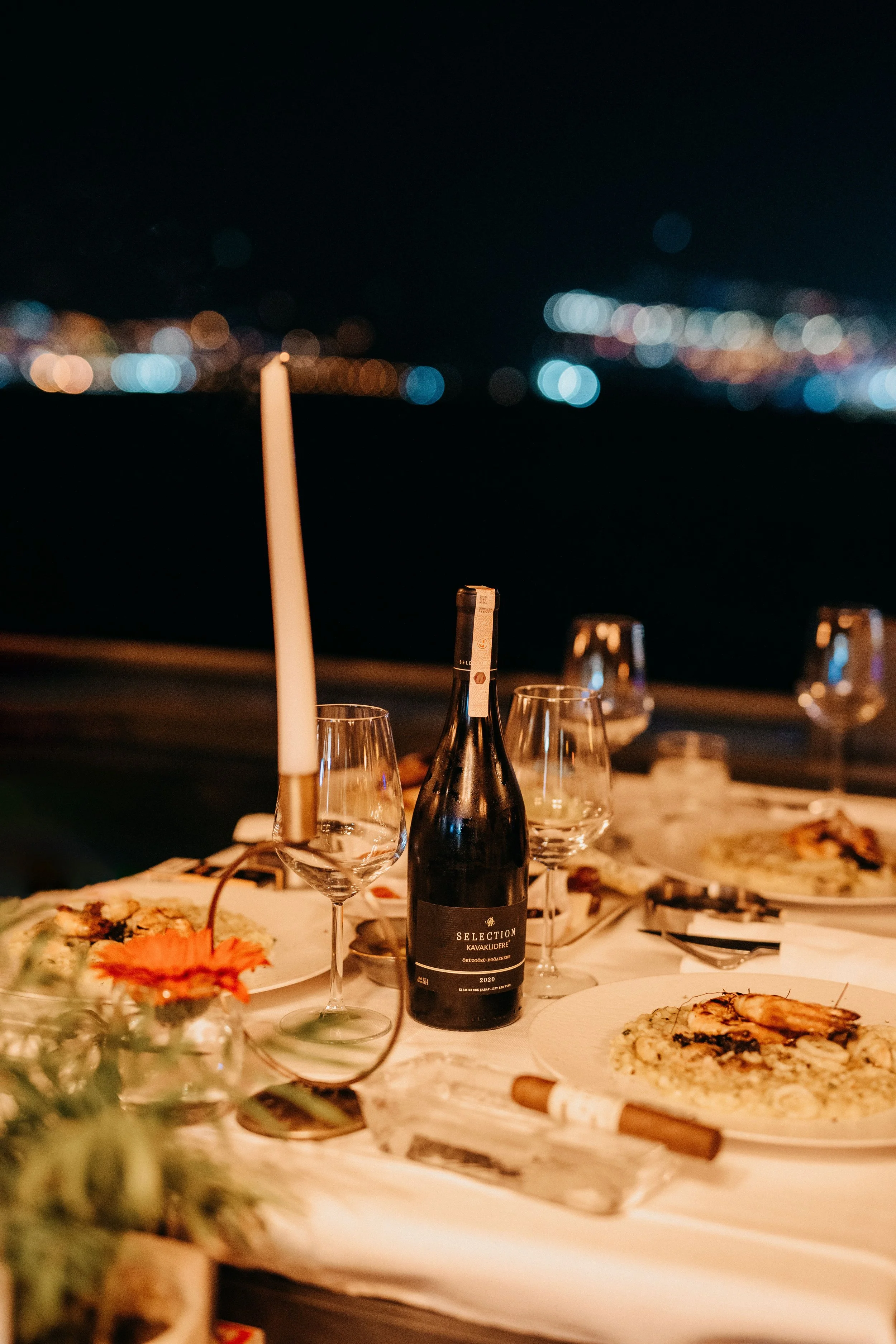 A dinner table at night with a bottle of red wine, empty wine glasses, plates of food, and blurred city lights in the background.