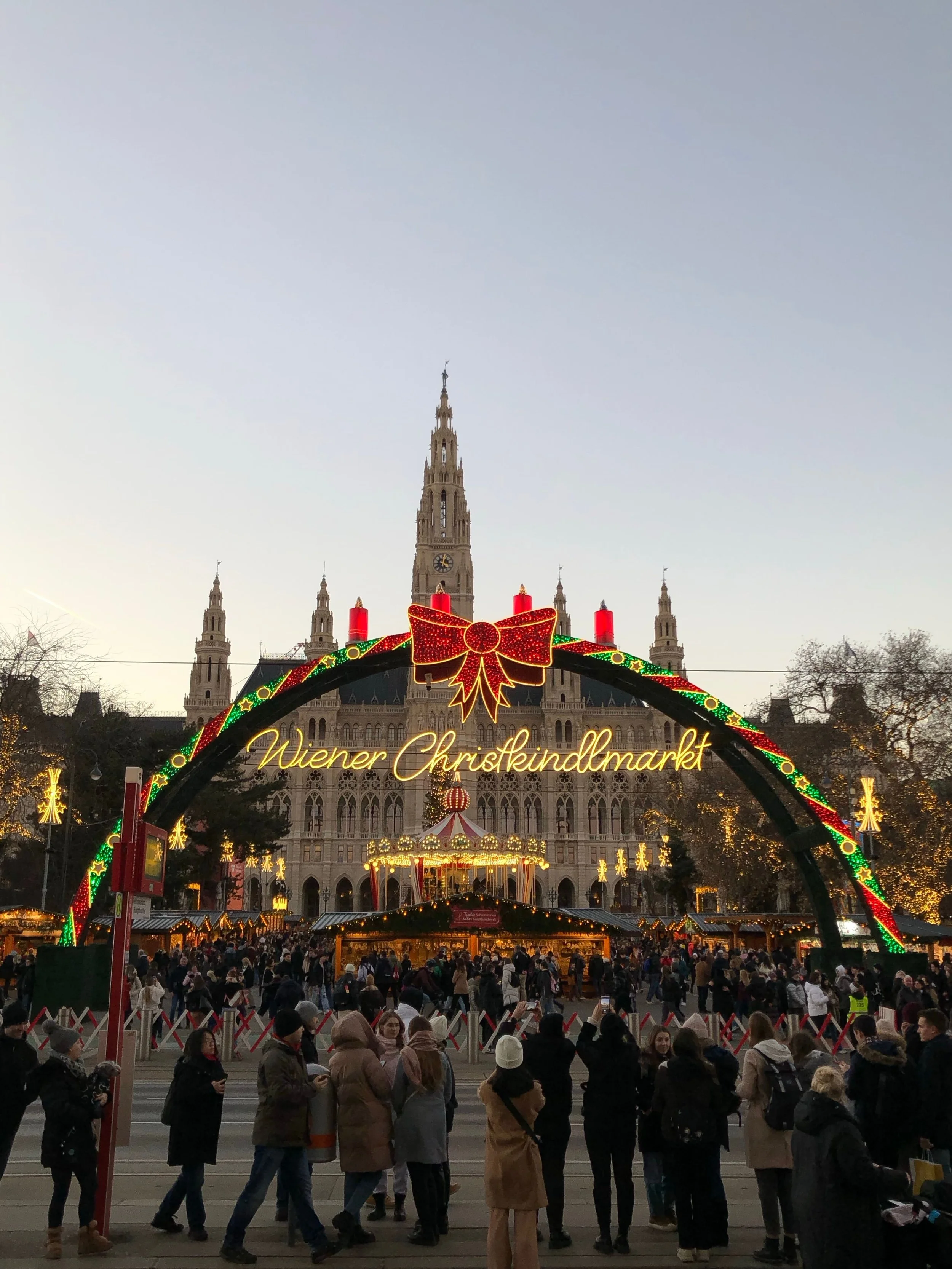 Christmas market in front of a historic building with a tall clock tower, decorated with lights, a large red bow, and festive ornaments, crowd of people shopping and walking.