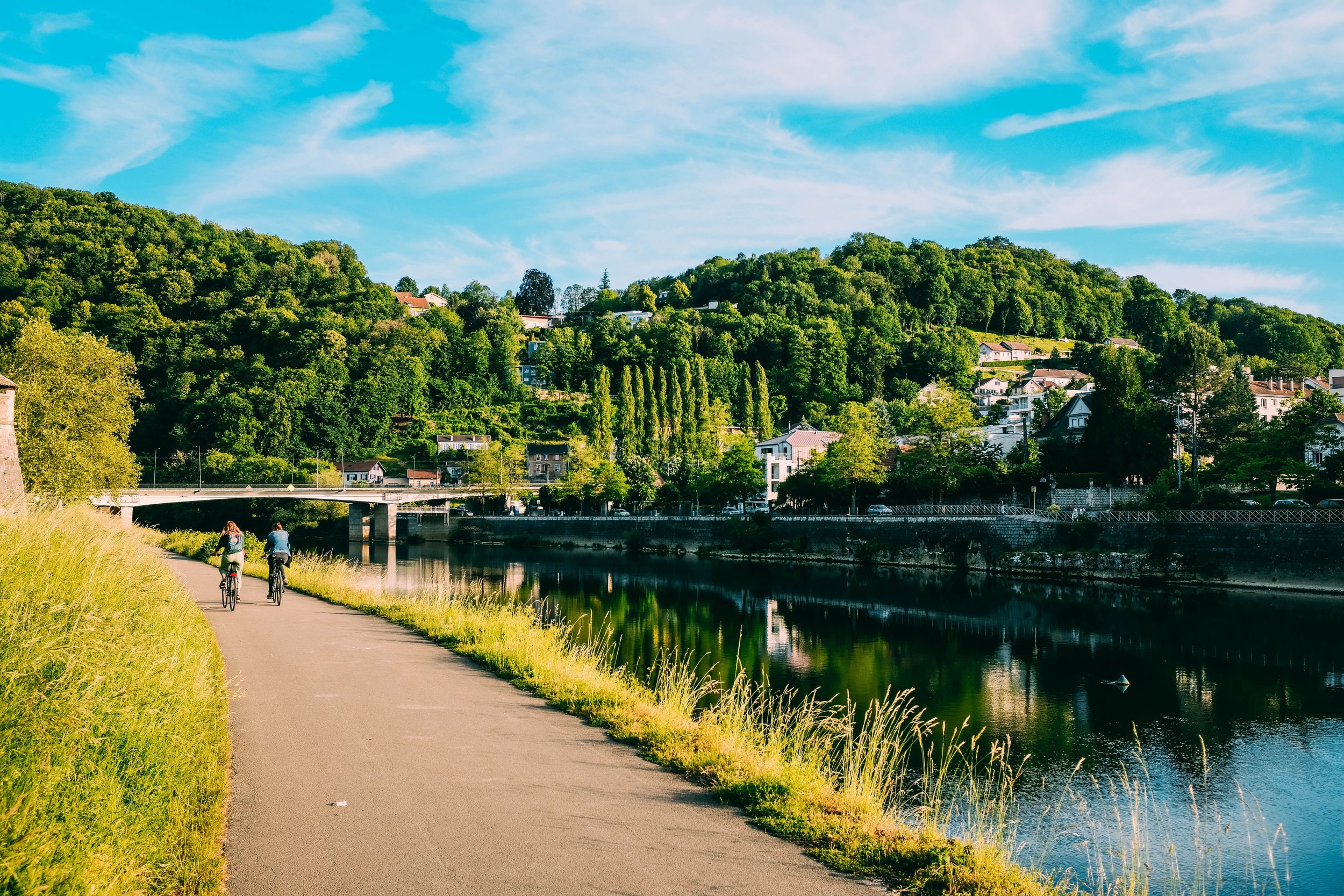 Two people riding bicycles along a paved pathway next to a calm river, with lush green hills and a small town in the background under a bright blue sky with white clouds.
