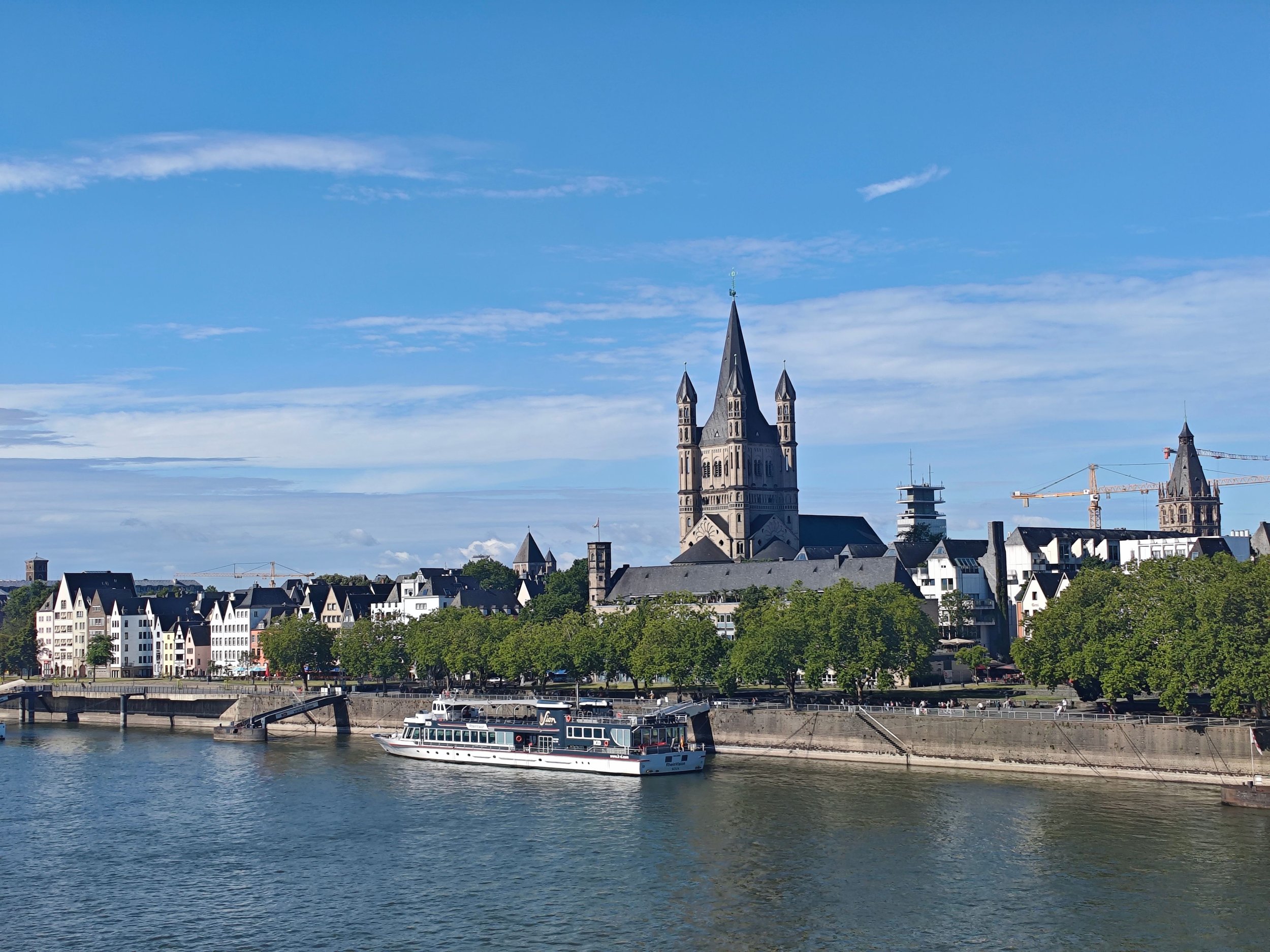 A city skyline with historic church and modern buildings by a river, boat in the water under a blue sky.