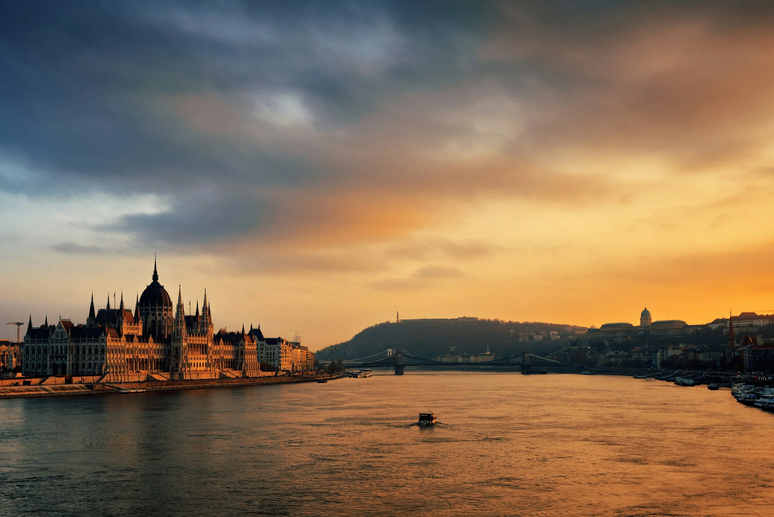 View of a city skyline along a river with historic buildings, bridges, and a hill with a monument at sunset.