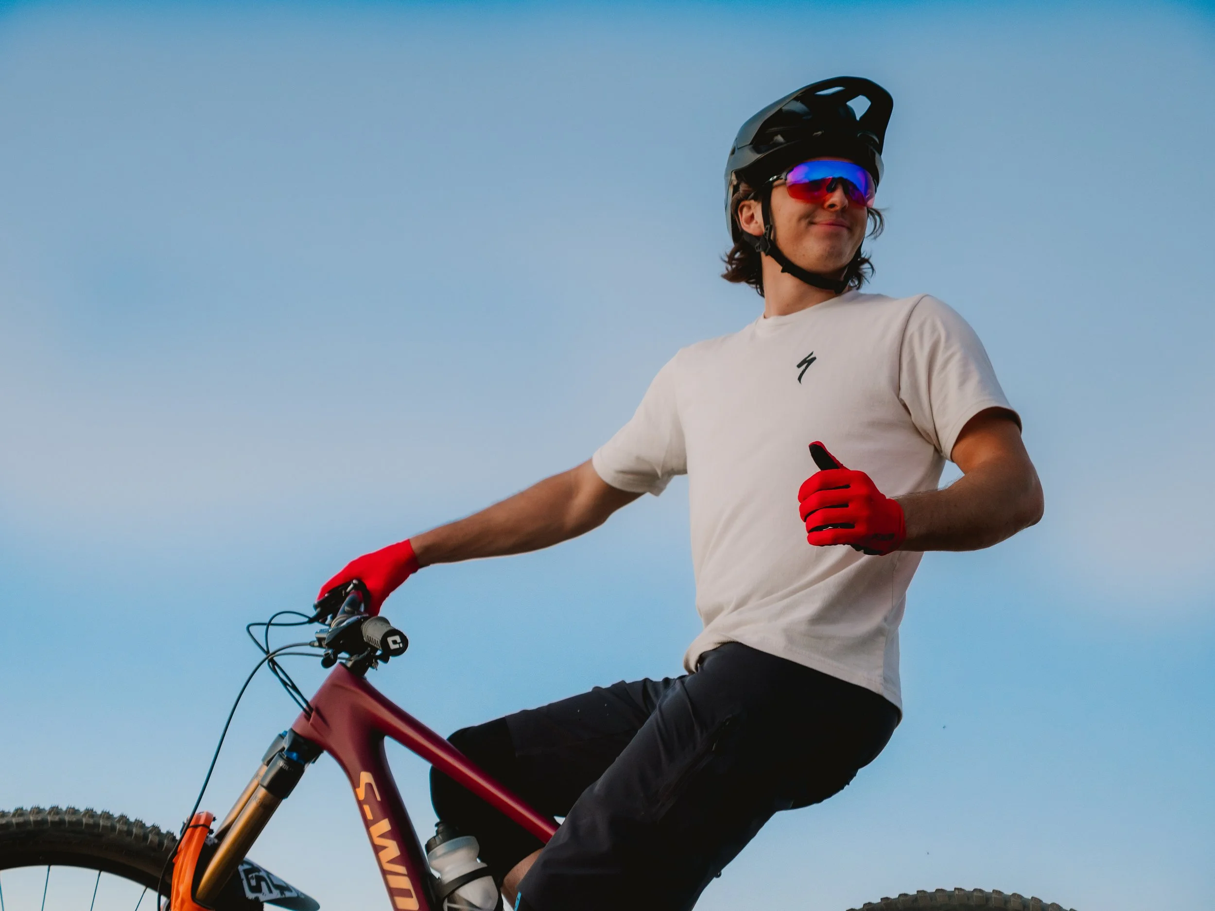 A person riding a mountain bike outdoors, wearing a black helmet, red-tinted sunglasses, a white t-shirt, and red gloves, with a clear blue sky in the background.