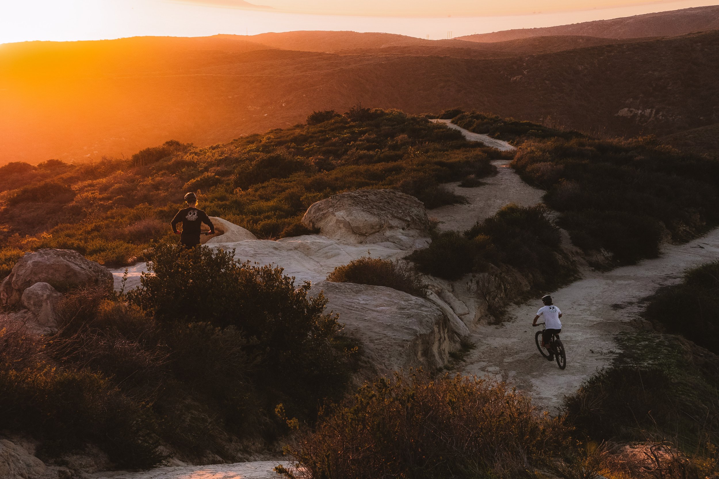 People riding bikes on a trail through hills at sunset with a person sitting on rocks in the foreground.
