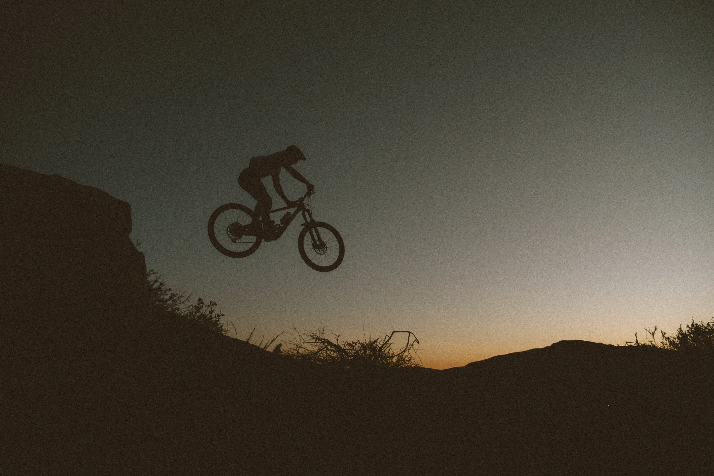 Silhouette of a person riding a mountain bike off a cliff at sunset or sunrise with a mountain landscape in the background.