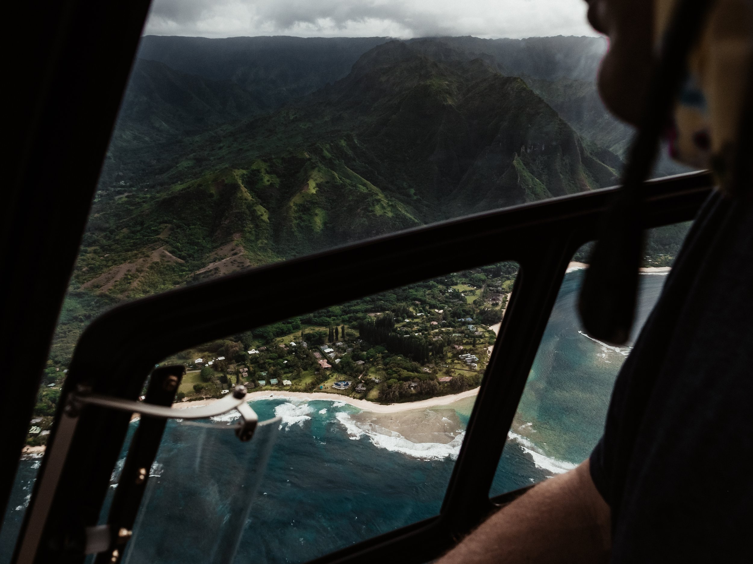 A helicopter flying over a lush green mountain range with a coastal area and houses below, cloudy sky overhead.