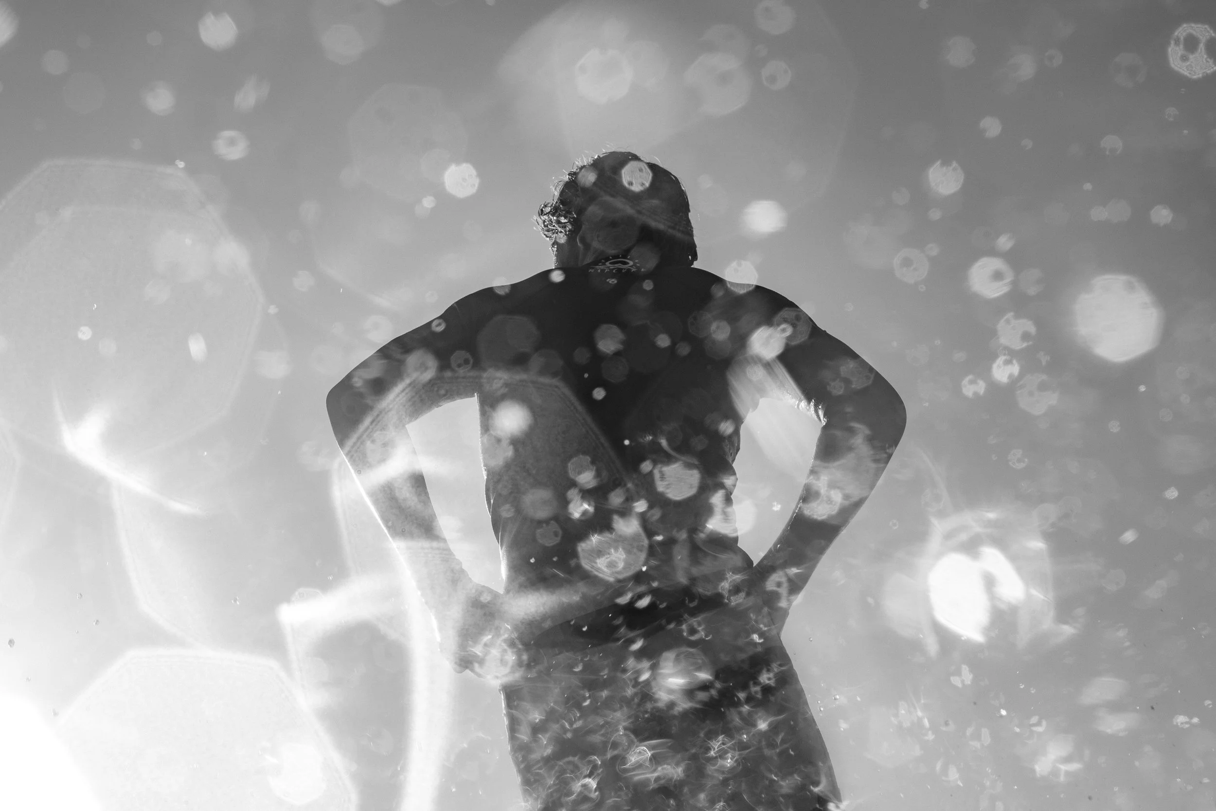 A person with curly hair wearing a wetsuit swims underwater in ocean water, surrounded by floating bubbles and sunlight filtering through the water.