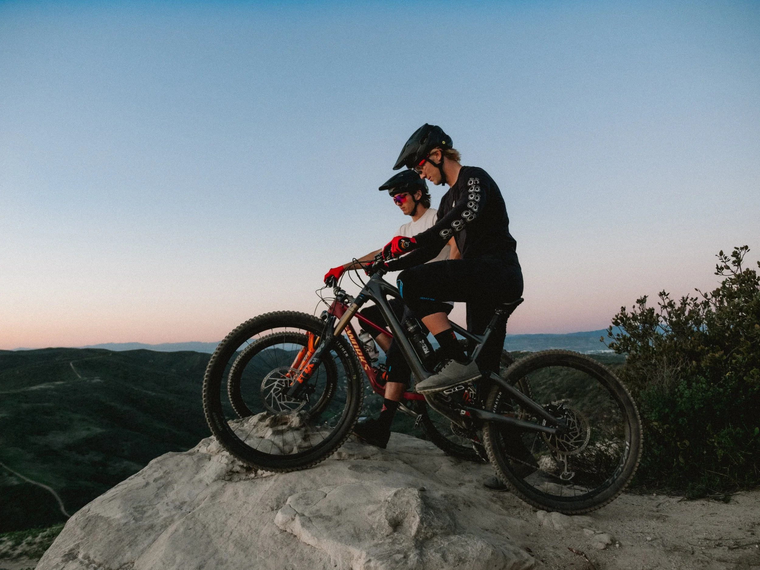 Two mountain bikers, wearing helmets and cycling gear, are riding their bikes on rocky terrain during dusk or dawn with a clear sky and distant mountains in the background.