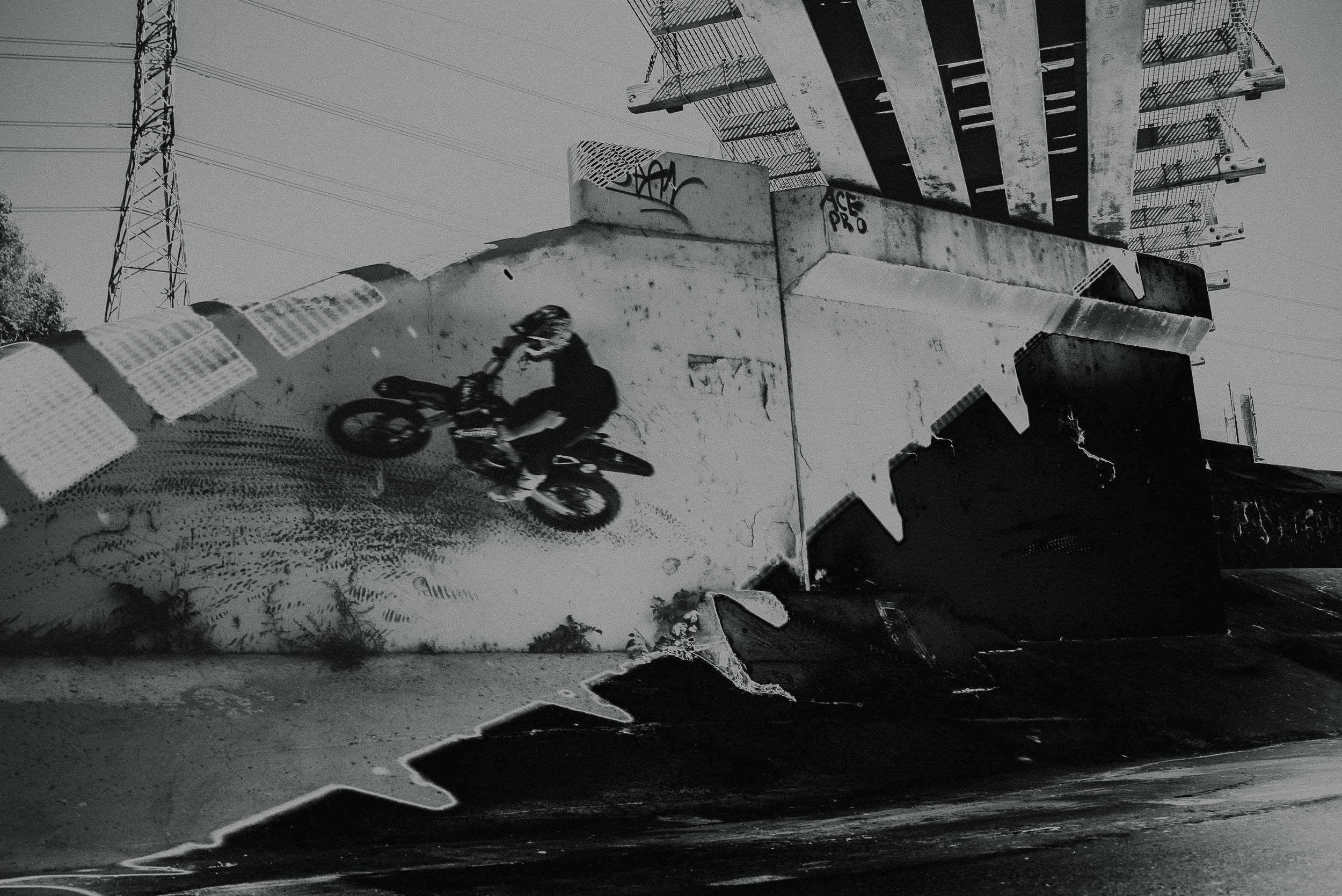 A monochrome photo of a skateboarder performing a trick at a skate park, with graffiti on the concrete structures and an industrial power line tower in the background.