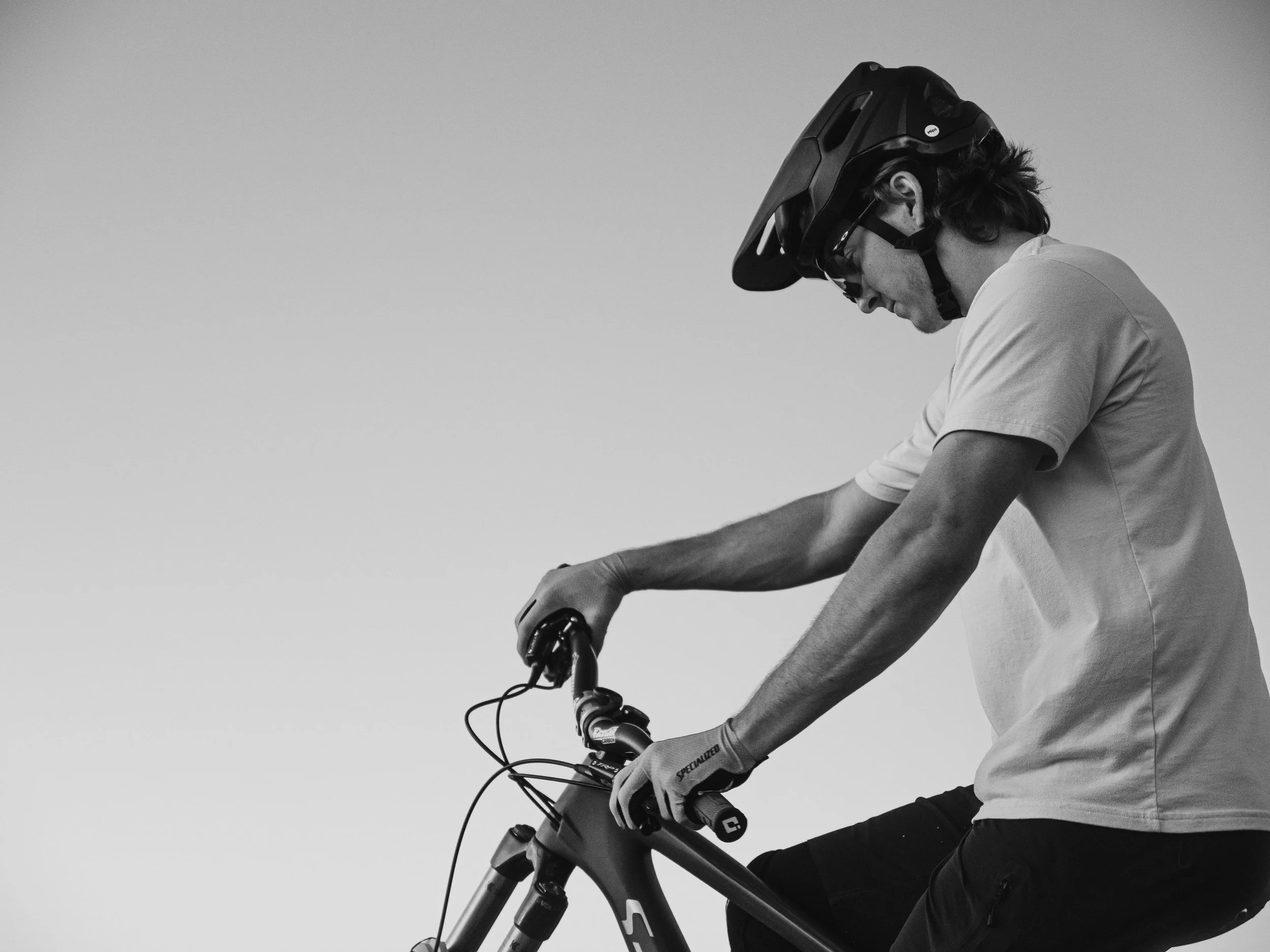 A person wearing a helmet and sunglasses riding a bicycle, looking downward with arms extended, against a plain background.