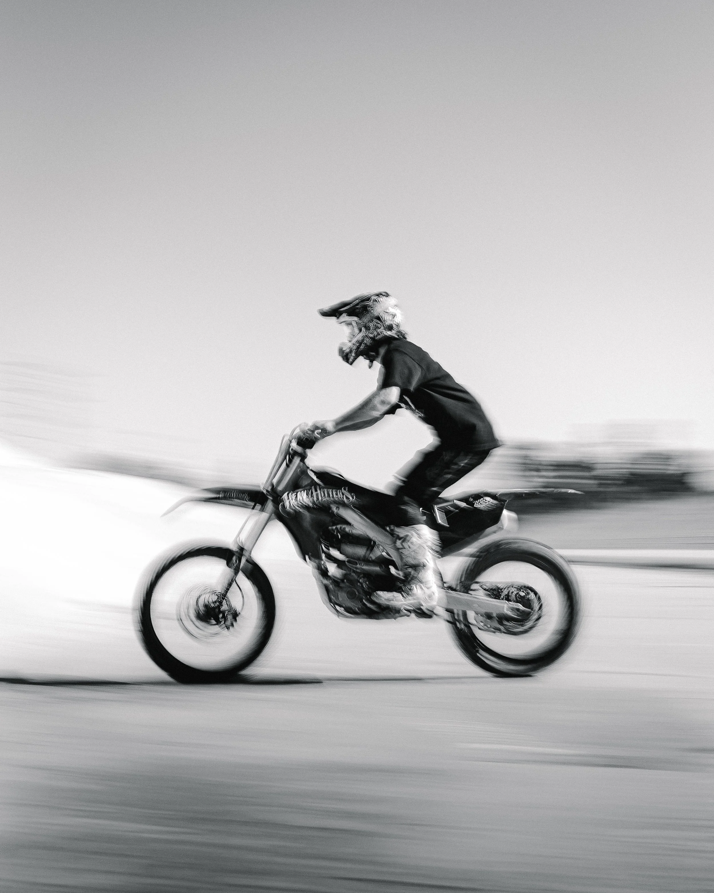 Black and white photo of a person riding a dirt bike at high speed, with motion blur showing the sense of movement against a blurred background.