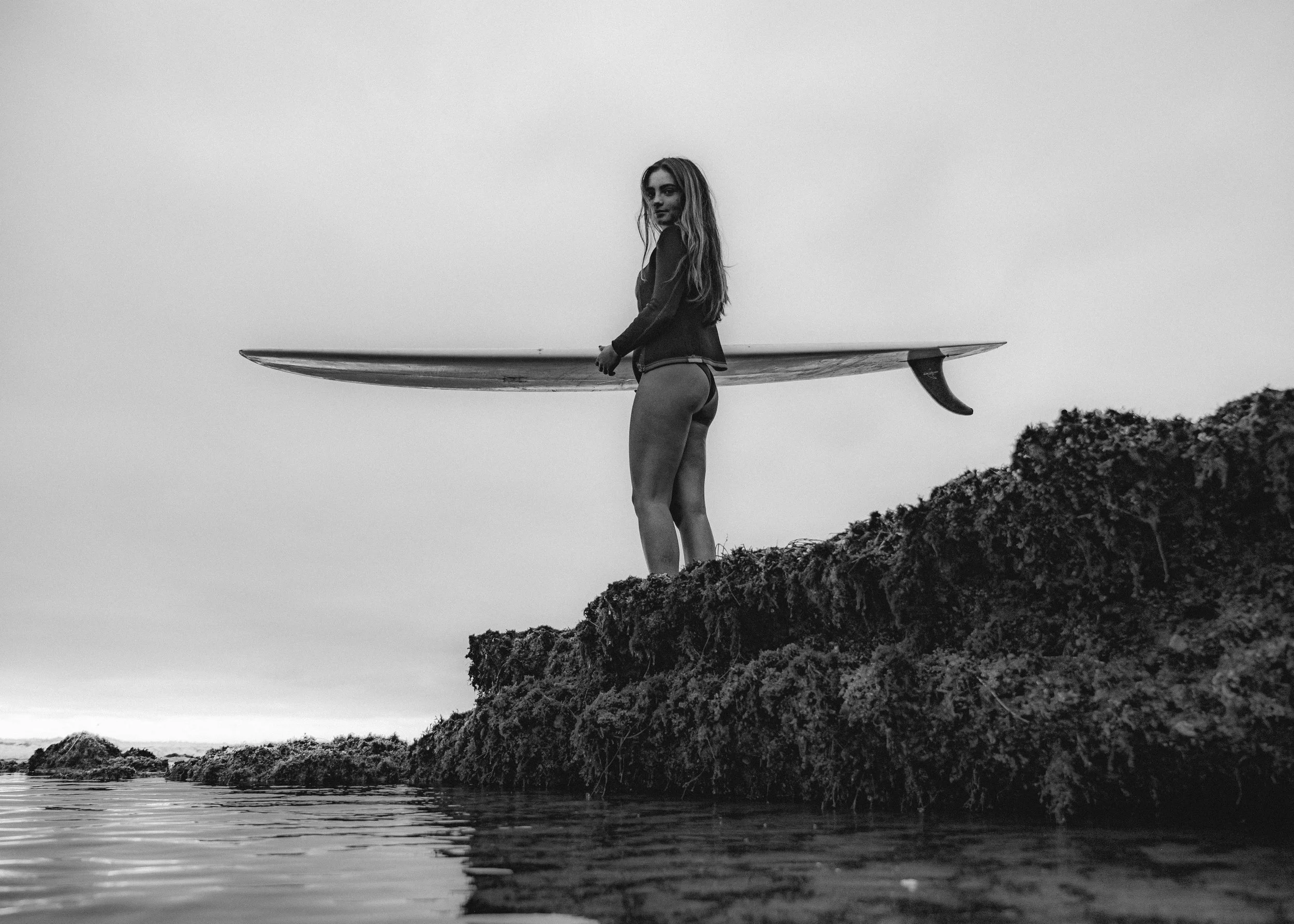 A woman holding a surfboard stands on a rocky shoreline, looking over her shoulder, with water and a cloudy sky in the background. The image is in black and white.
