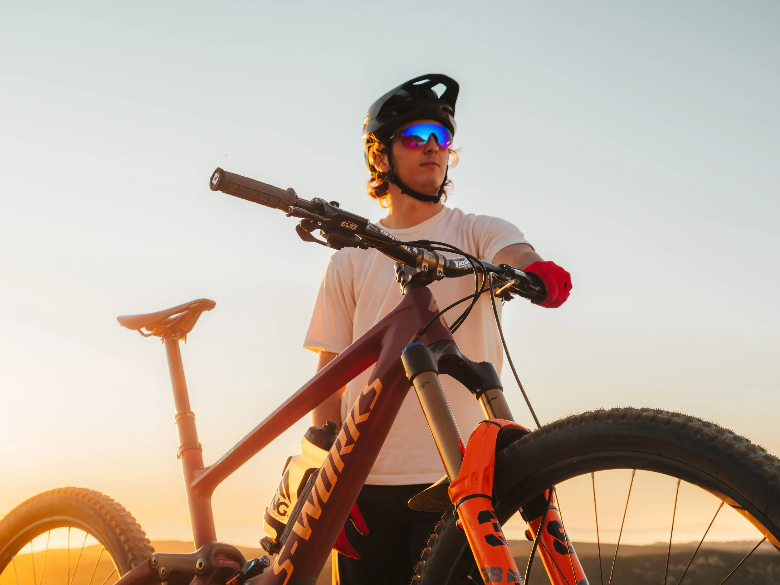 A person wearing a black helmet, colorful reflective sunglasses, and a beige shirt, standing with a mountain bike during sunset or sunrise.