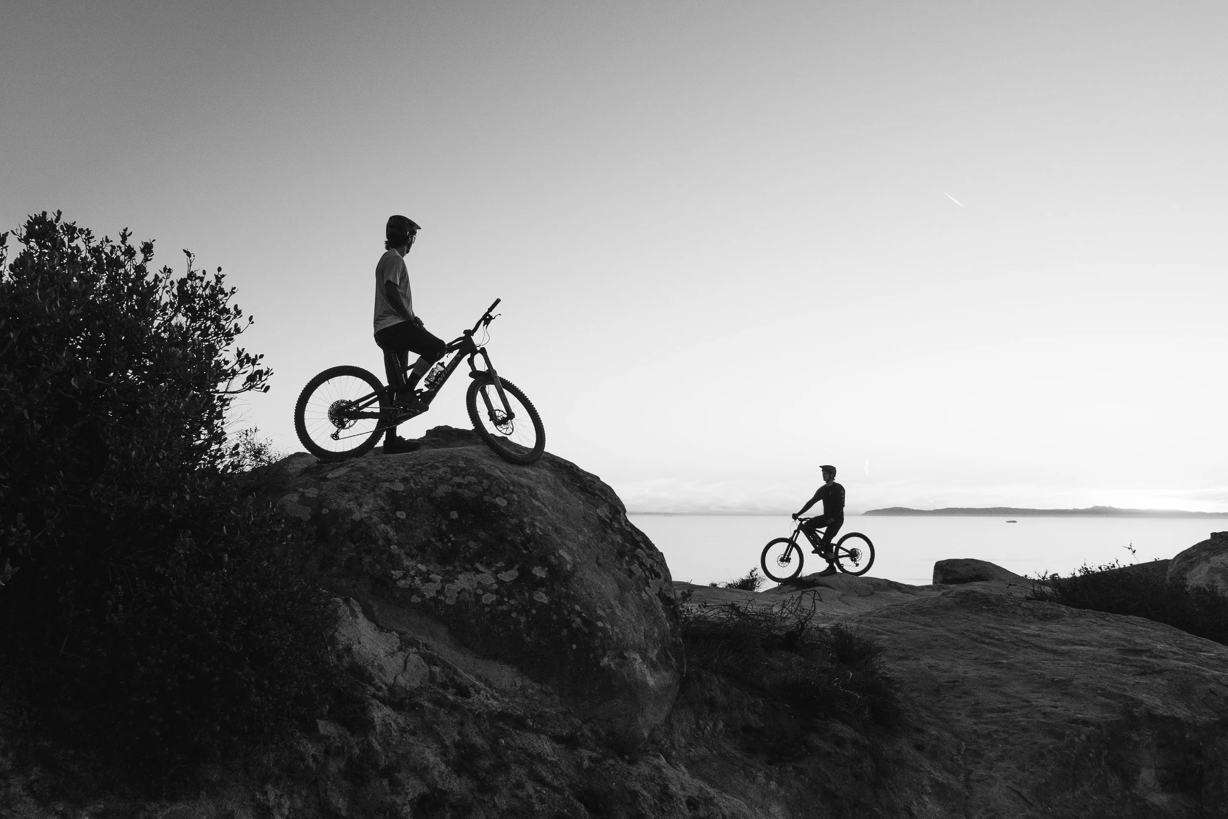 Two people with bicycles on rocky terrain near a body of water during sunset, with one person standing on a rock and the other riding a bike.