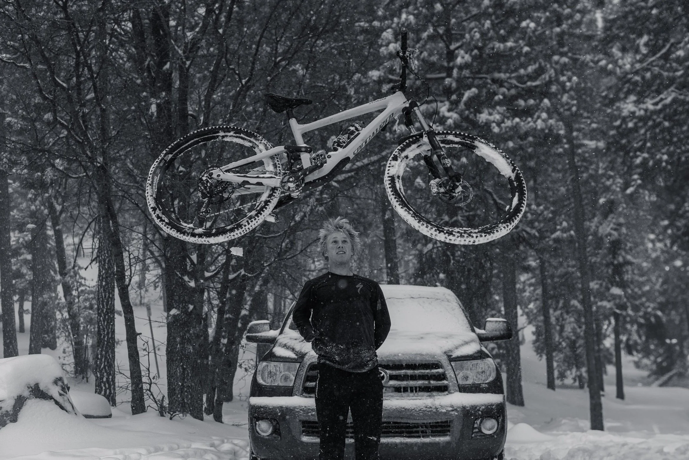 A man standing in a snowy outdoor setting with a car behind him and a mountain bike mid-air above his head during snowfall.