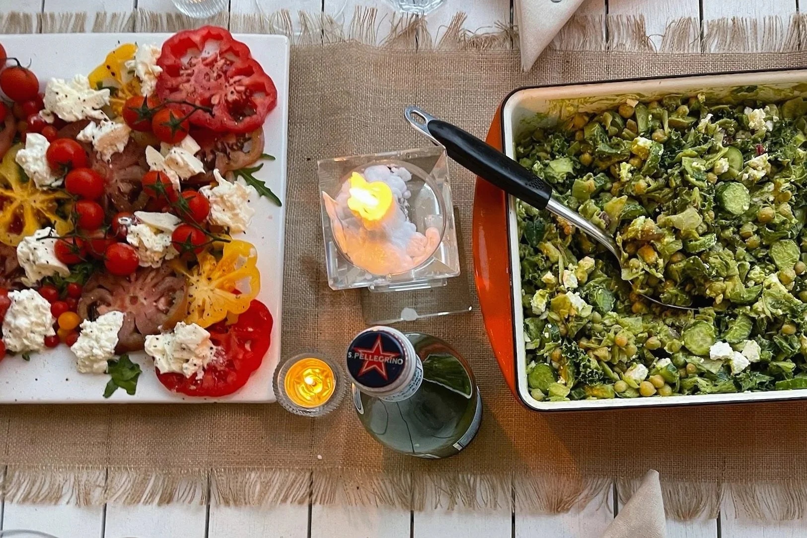 A table with a colorful salad with cherry tomatoes, mozzarella, and sliced vegetables on a white plate, a large tray of chopped green vegetables with cheese, a lit candle, and a bottle of beer.