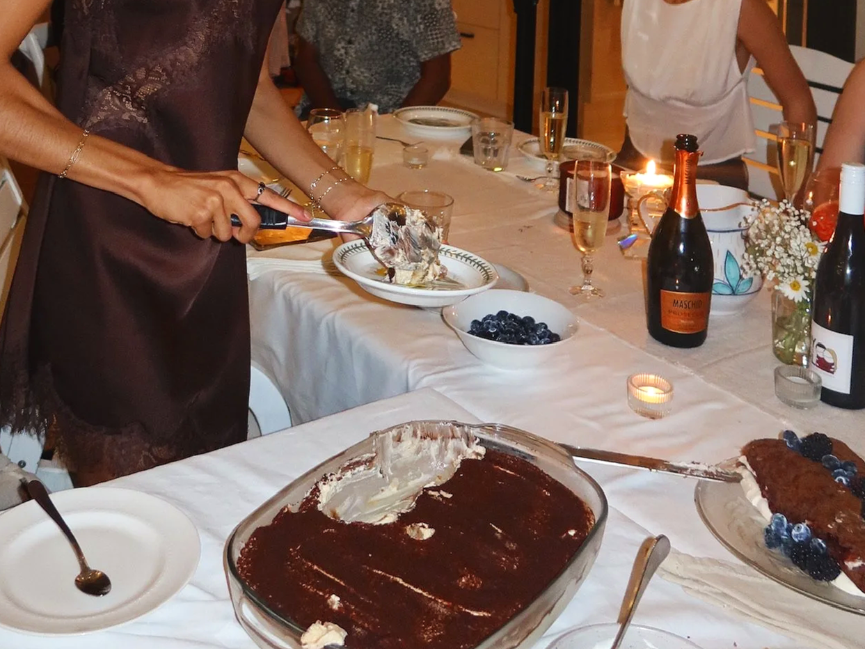 A woman serving dessert at a dinner party with a large chocolate cake topped with blueberries, bottles of sparkling wine, and various glasses on a table decorated with flowers and candles.