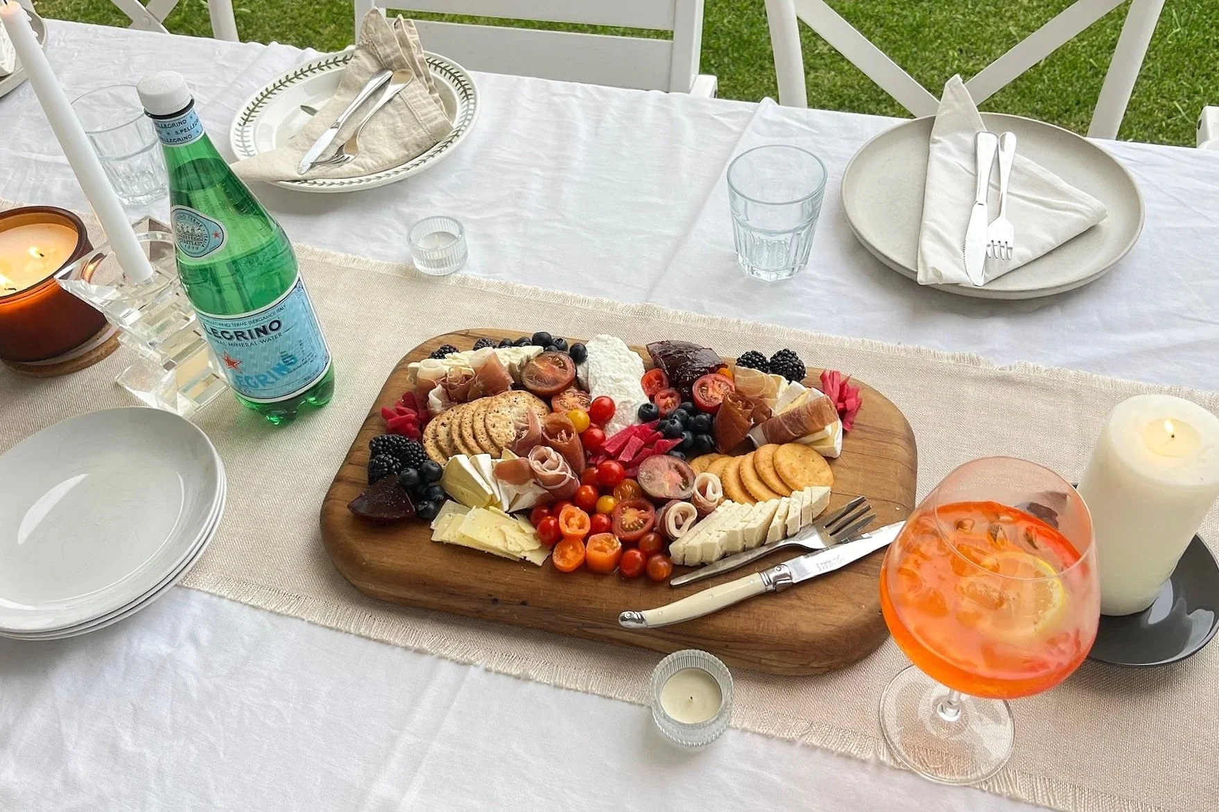 A table set for an outdoor meal featuring a wooden platter with various cheeses, fruits, crackers, and charcuterie. The table has white plates, glasses, cutlery, and candles, with a green glass bottle of sparkling water and a large glass of orange-colored drink with slices of citrus. The background shows a grassy area and white chairs.