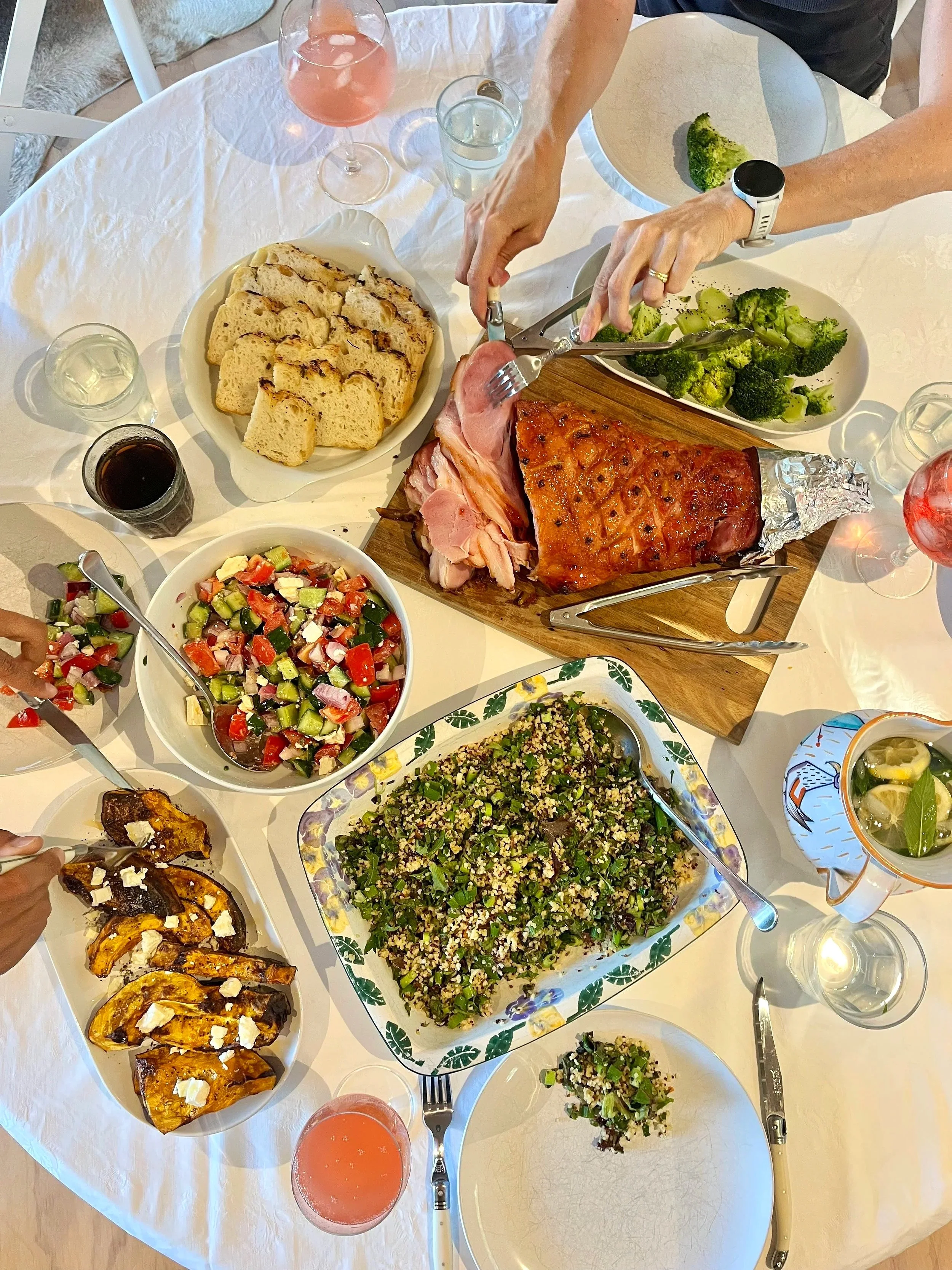 A table set with various dishes including sliced bread, roasted meat, broccoli, a colorful vegetable salad, roasted eggplant, and two glasses of pink and clear drinks. There are utensils and napkins on the table.