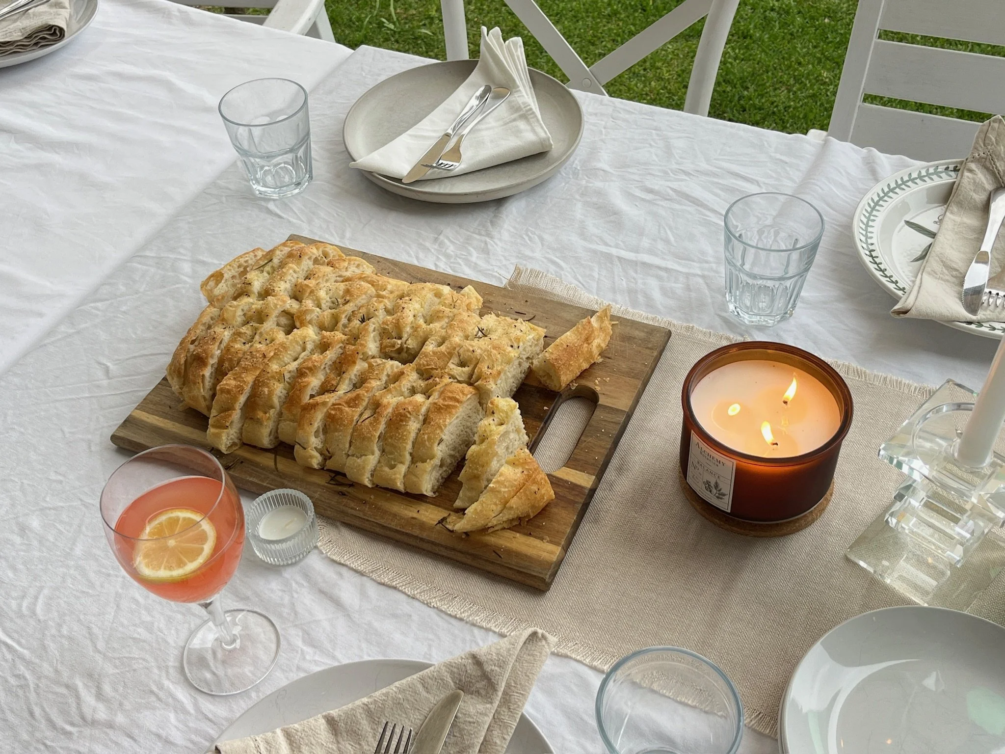 A white outdoor dining table set with clear glasses, plates with patterned green border, forks, knives, and cloth napkins. There is a loaf of bread with a lattice top on a wooden cutting board, a lit candle in a glass holder, and a glass of pink lemonade with lemon slices. Chairs with white slats are around the table, and green grass is visible in the background.