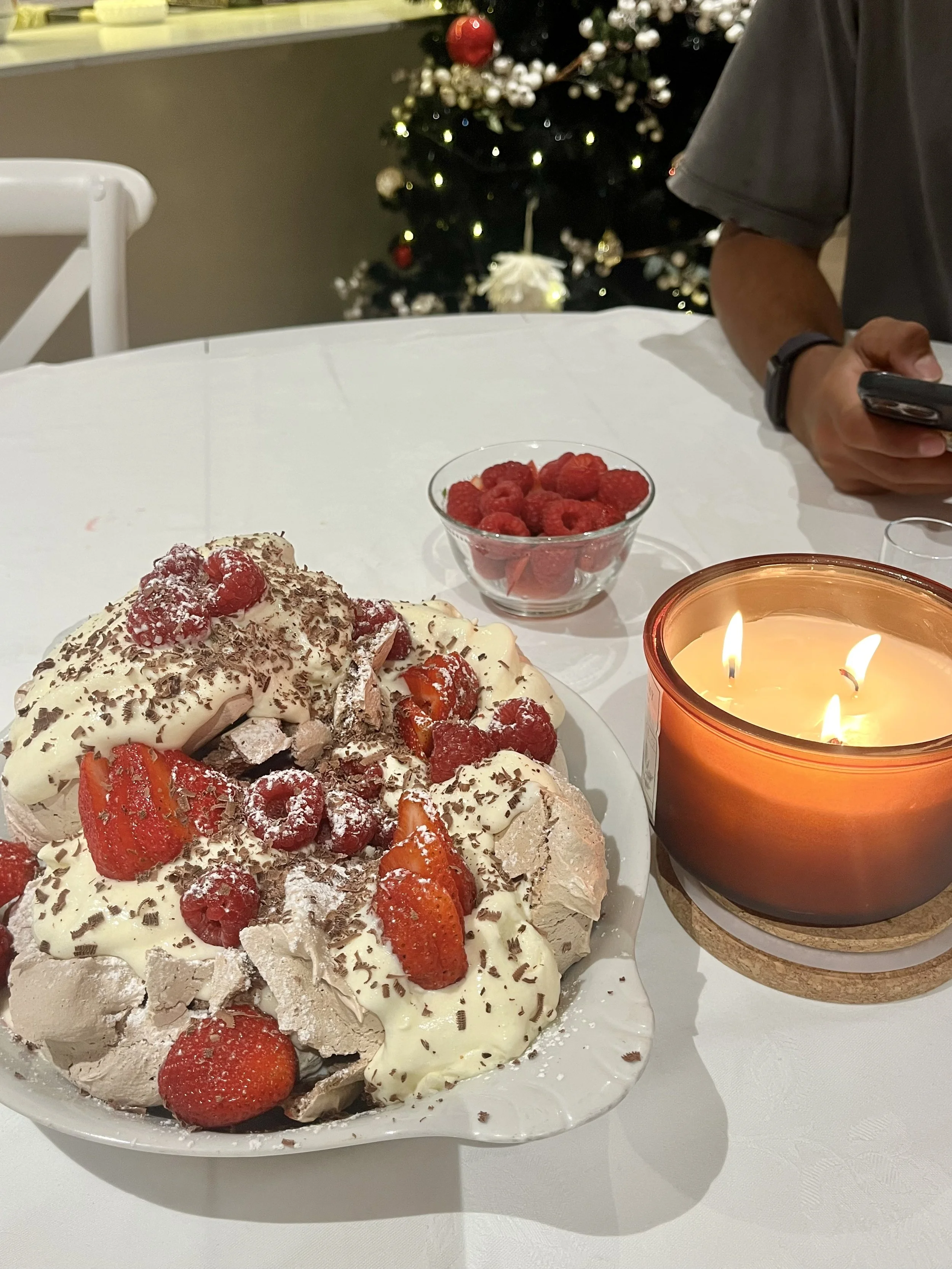 A dessert topped with strawberries, raspberries, chocolate shavings, and whipped cream on a white plate, a bowl of raspberries, a lit candle, and a Christmas tree in the background.