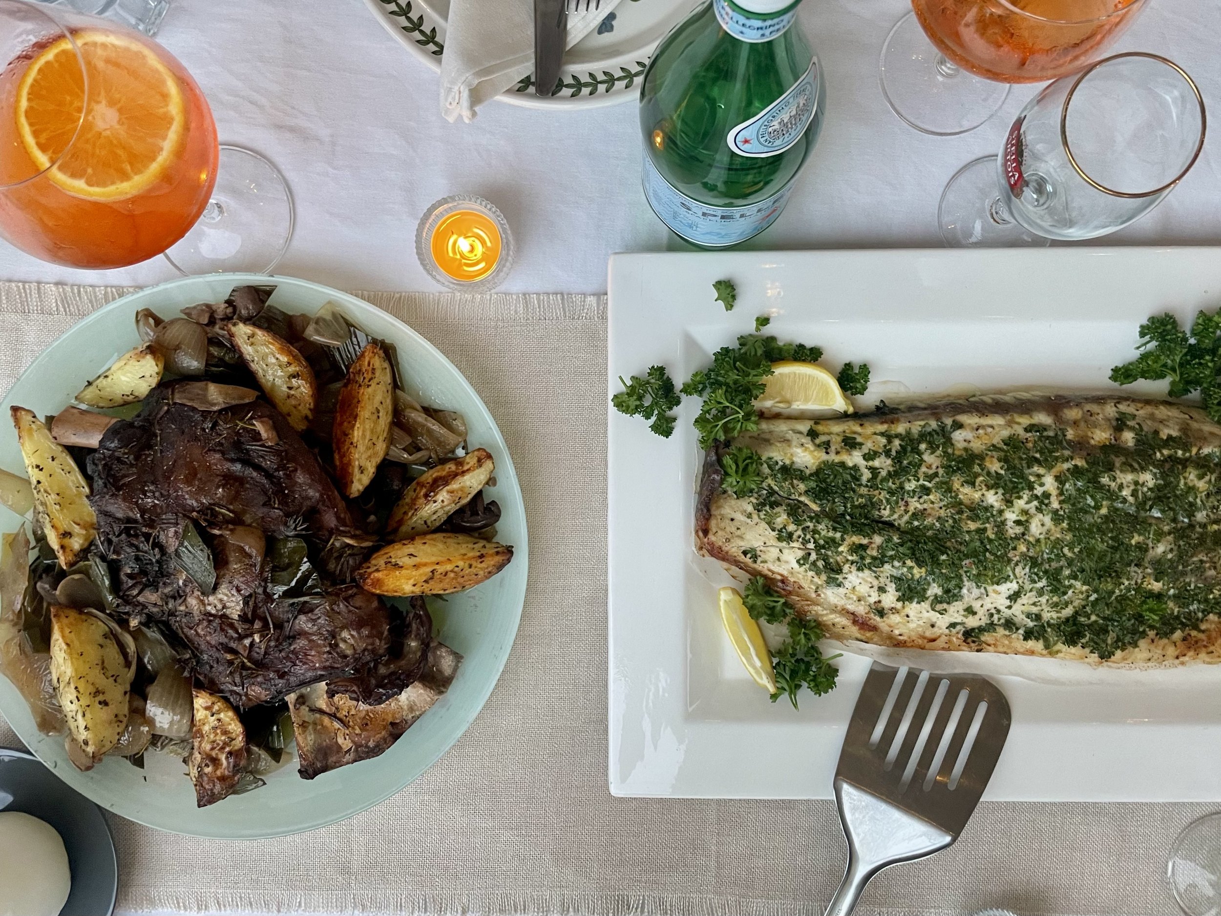A table with seafood and drinks. There is a whole fish garnished with herbs, lemon, and greens on a rectangular white plate. A bowl with roasted potatoes and vegetables, and another bowl with roasted vegetables are on the table. There are two glasses of rosé wine and a bottle of San Pellegrino sparkling water. A small candle is lit on the table.