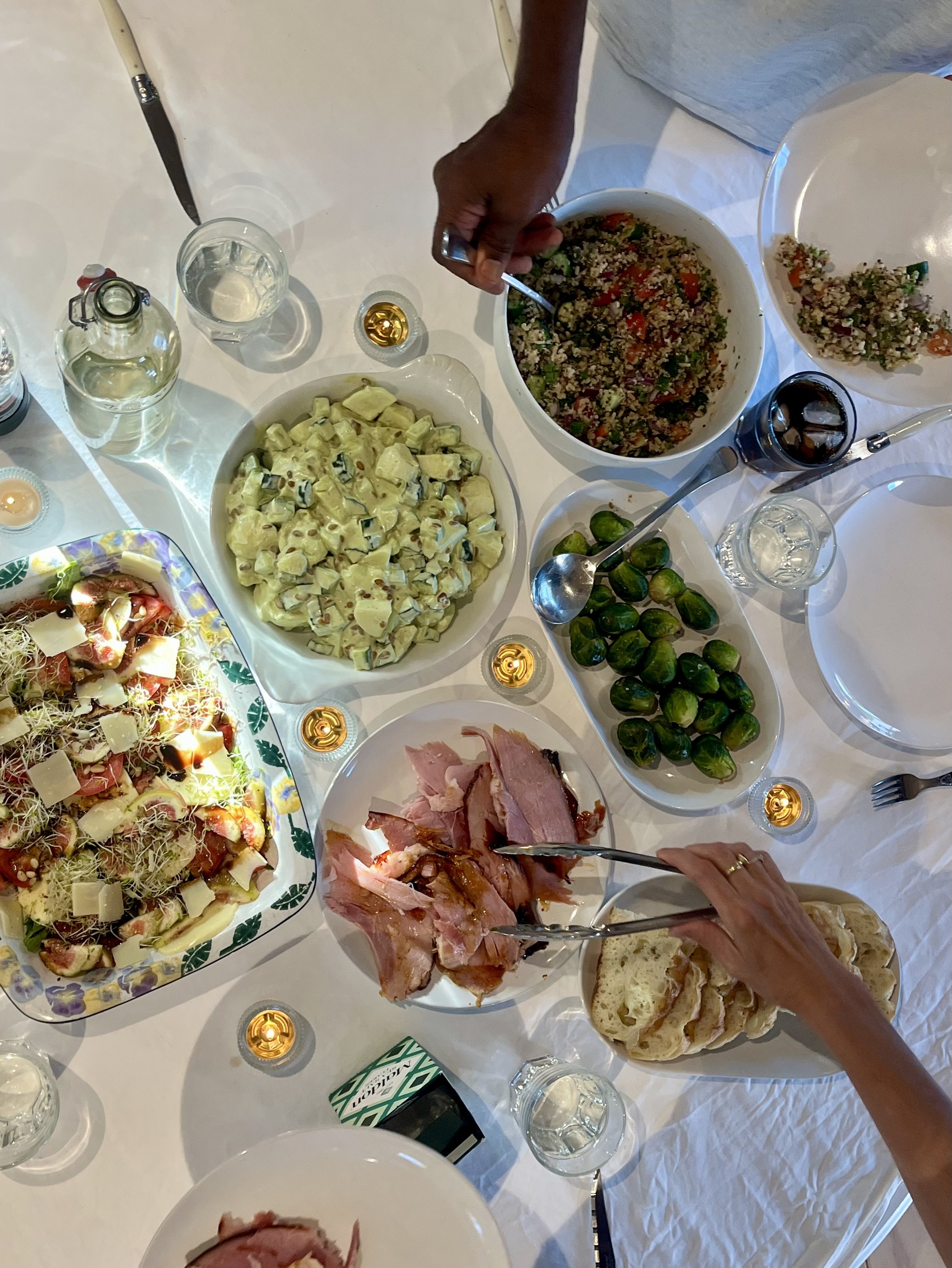 A table with various dishes, including salad, potato salad, sliced meats, bread, and drinks, with a person serving food.