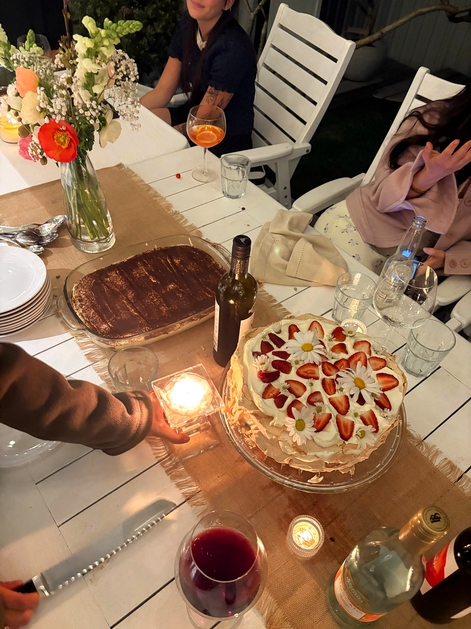 A birthday celebration outdoors at night, with a table decorated with a large strawberry cake topped with strawberries and flowers, a bottle of wine, glasses of red wine, a glass of orange drink, flowers, candles, and people sitting around the table.