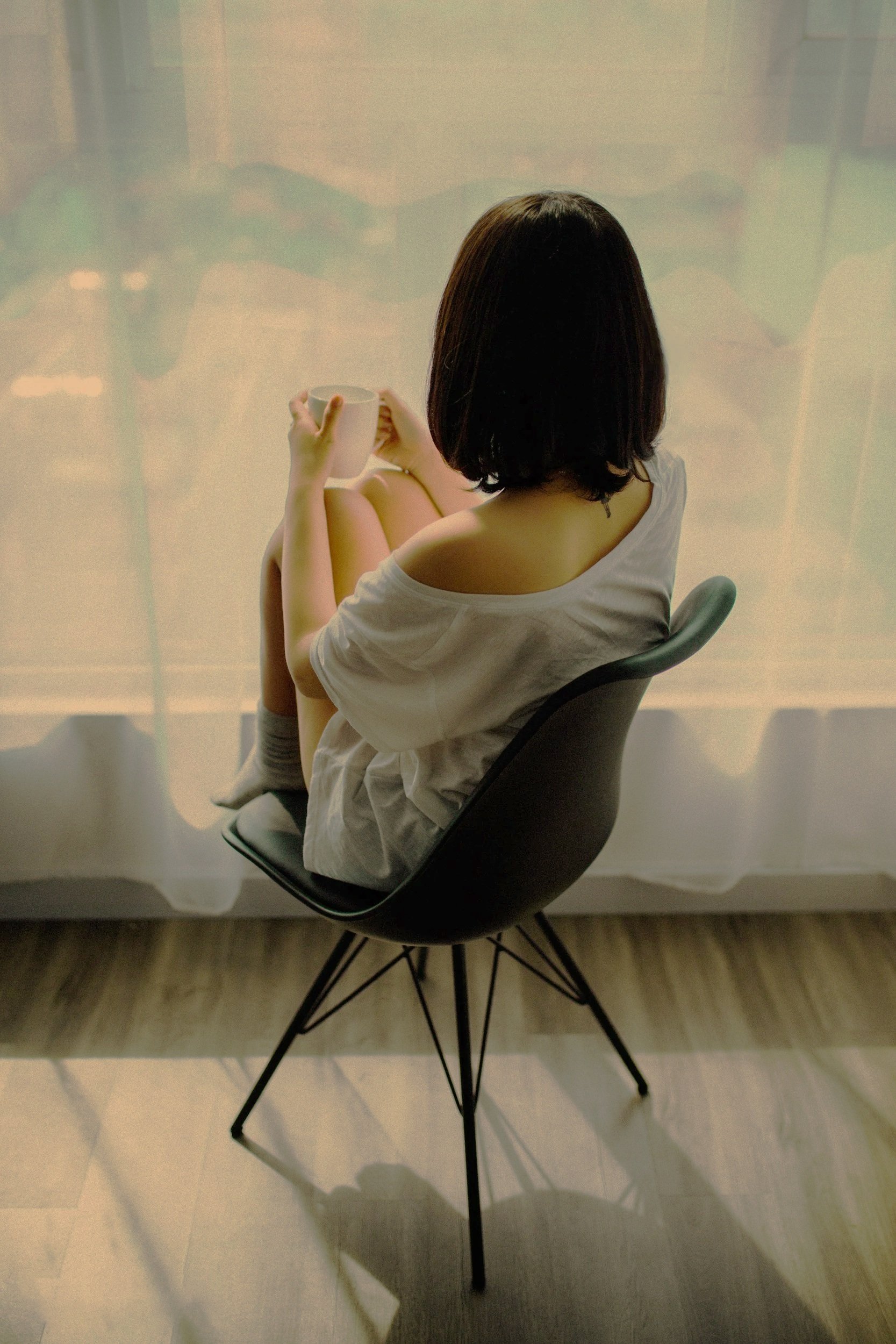 A woman sitting in a modern black chair, viewed from behind, holding a white mug, with a sheer curtain in the background and soft lighting.