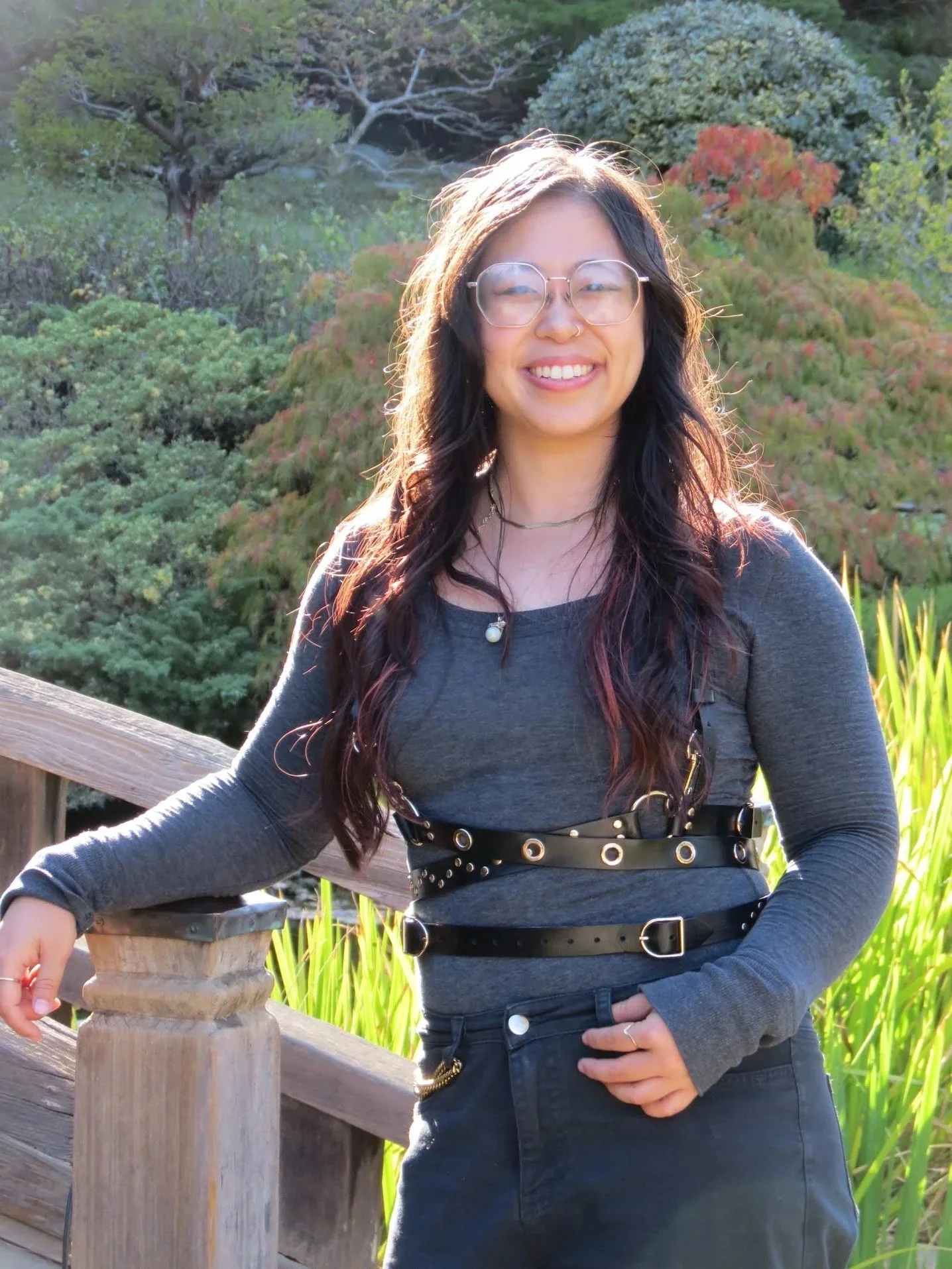 A young woman with long dark hair and glasses standing outdoors near a wooden railing, smiling at the camera, with green foliage and trees in the background.