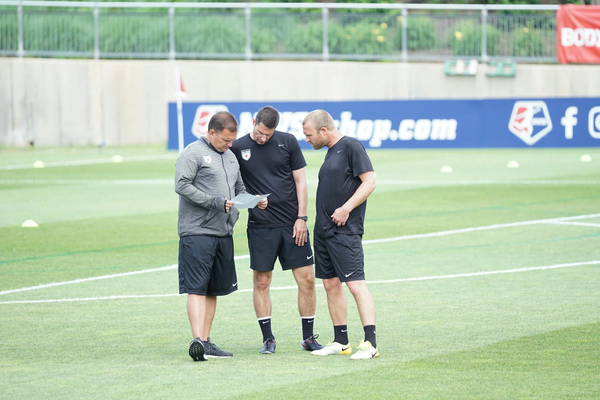 Three men standing on a green soccer field, looking at a paper. Two are wearing black athletic shirts and shorts, the third in a gray jacket and dark pants. There is a red and white soccer ball on the ground near their feet.