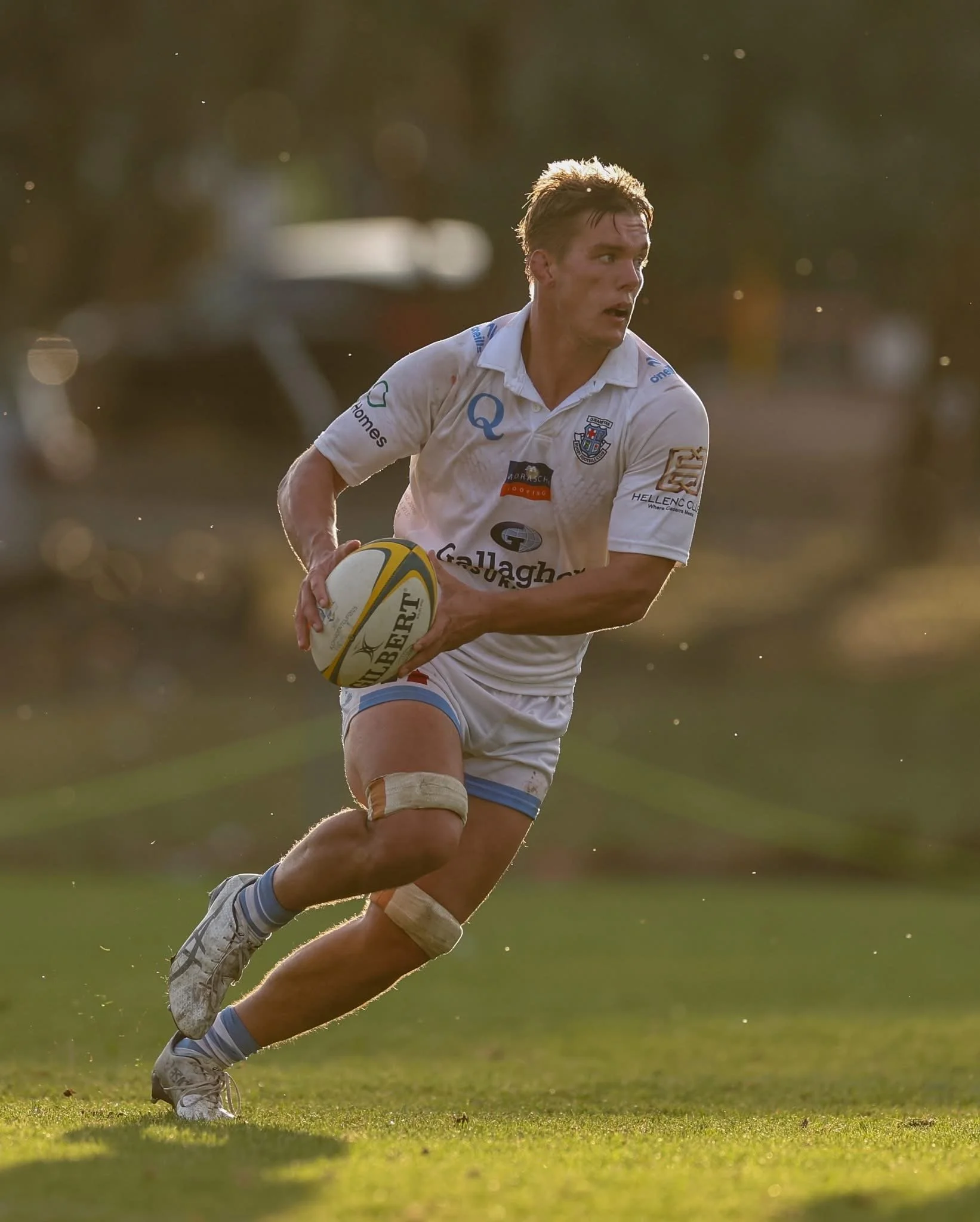 A rugby player running on the field holding a yellow and white rugby ball, wearing a white jersey with logos, shorts, and athletic shoes, with sunlight and blurred background.