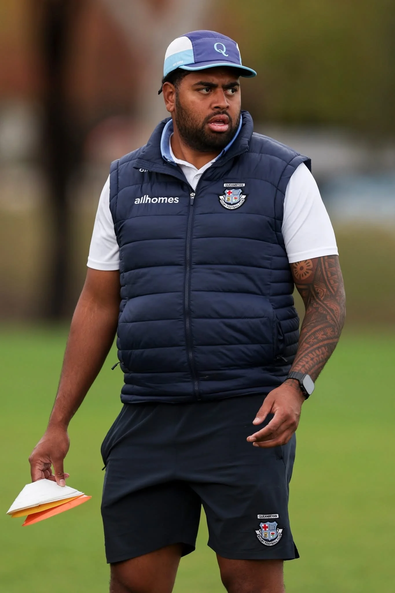 A man with a beard and tattoos on his arm, wearing a blue and white cap, a navy vest with the logo of Queanbeyan Rugby Football Club and the word 'allhomes,' a white polo shirt, and black shorts with the same logo, stands outdoors on a grassy field, holding a stack of papers or clipboards, looking to his left with a focused expression.