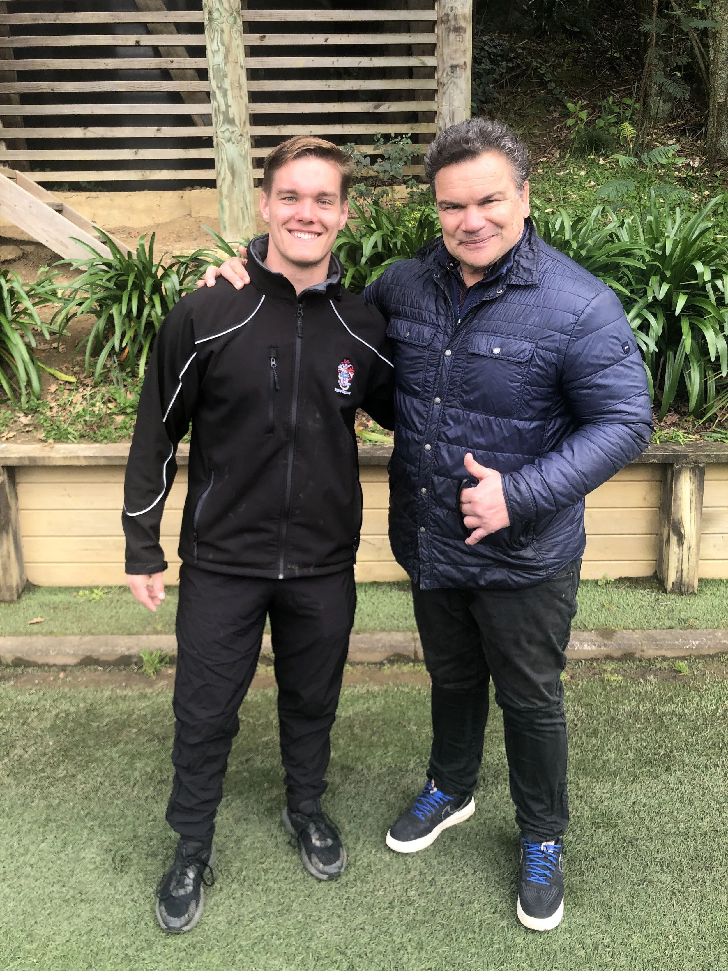 Two men standing outdoors on grass, smiling at the camera, one in a black jacket and the other in a navy blue quilted jacket, with greenery and a wooden fence behind them.