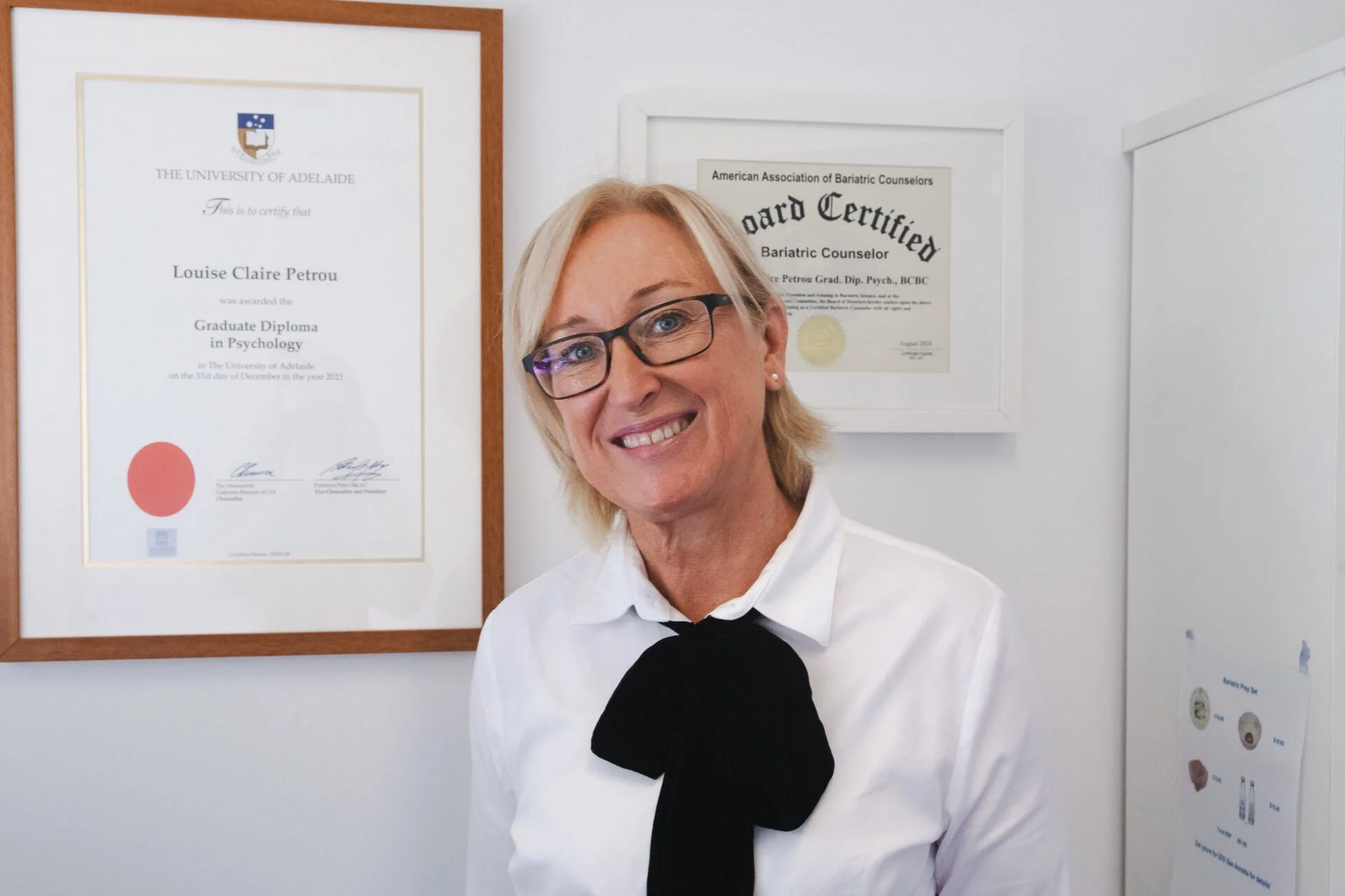 A woman with blonde hair, glasses, and a white shirt standing in front of framed diplomas on a white wall.
