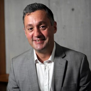 Professional man with short dark hair, wearing a gray suit and white shirt, smiling against a gray wall background.