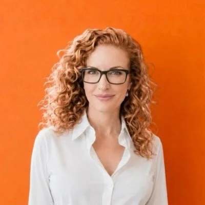 A woman with curly red hair and glasses smiling against an orange background.