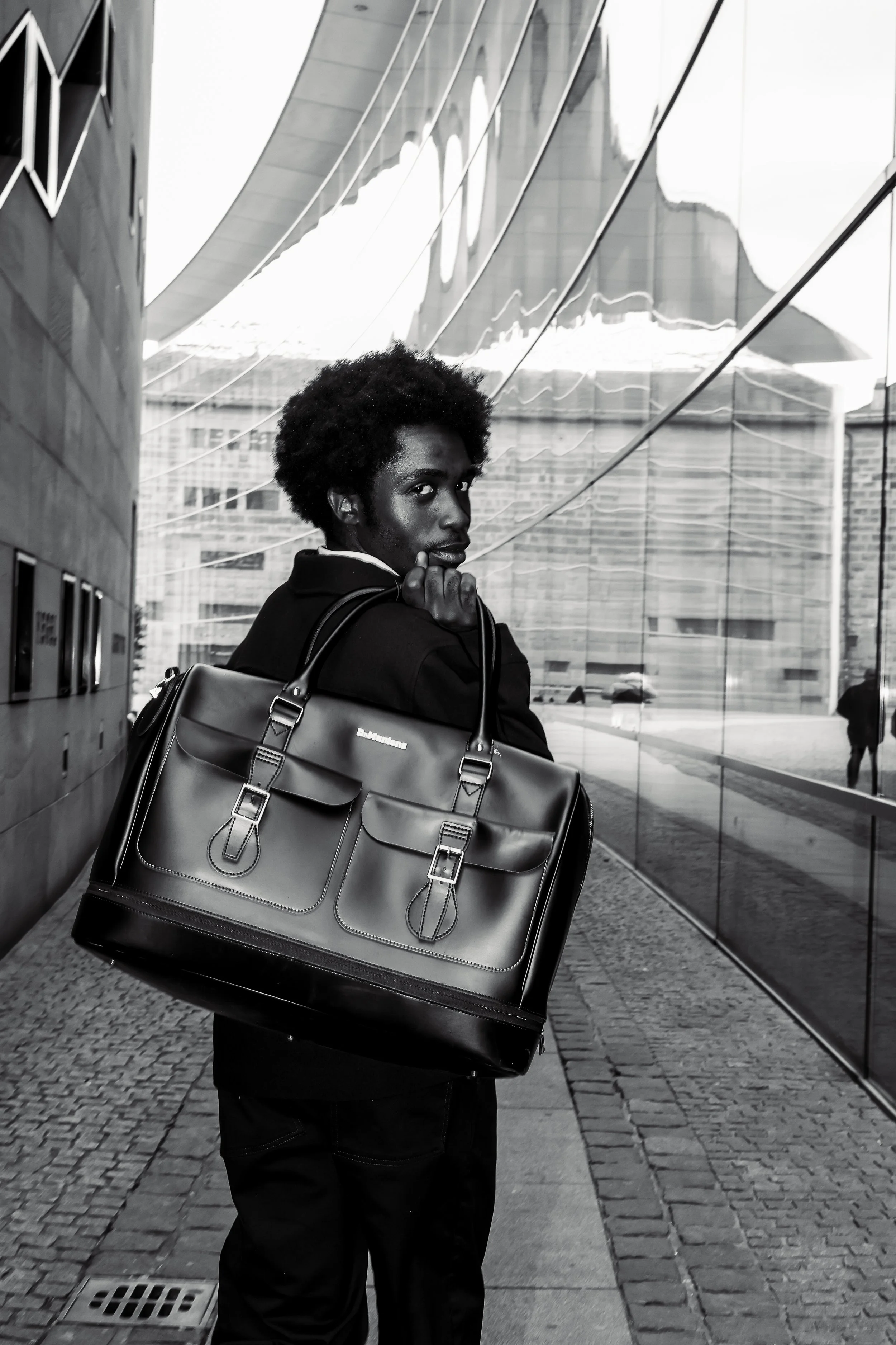 Ein Mann mit dunkler Haut und Afro-Haar trägt einen schwarzen Mantel und eine große braune Aktentasche, die er über seine Schulter hängt. Er steht vor einer reflektierenden Glasfassade eines Gebäudes, in dem sich seine Silhouette spiegelt.