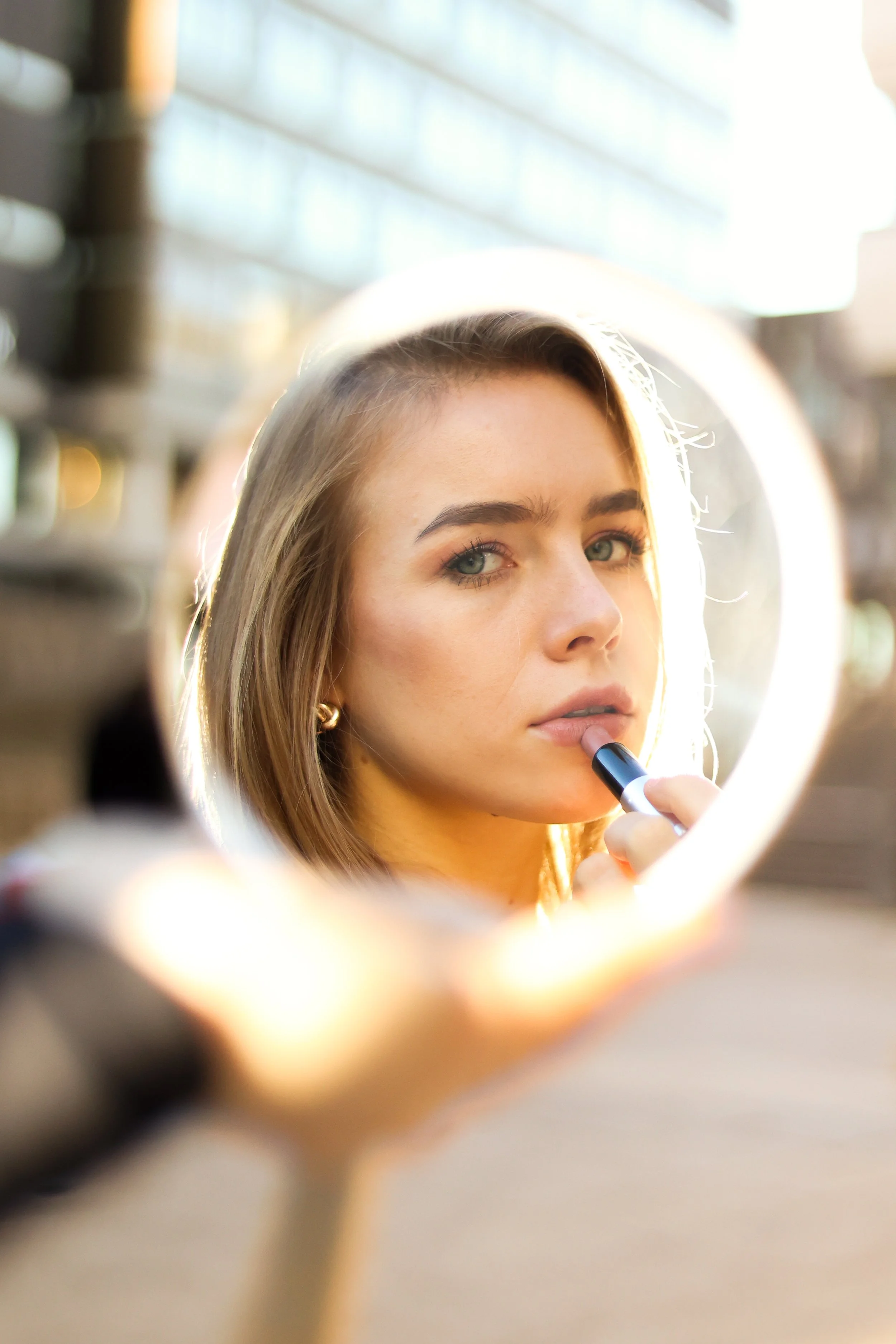 Ein junges Frau, die in einem Spiegel Make-up aufträgt, sichtbar durch den Rundspiegel, mit Hintergrundbeleuchtung in einem Raum.