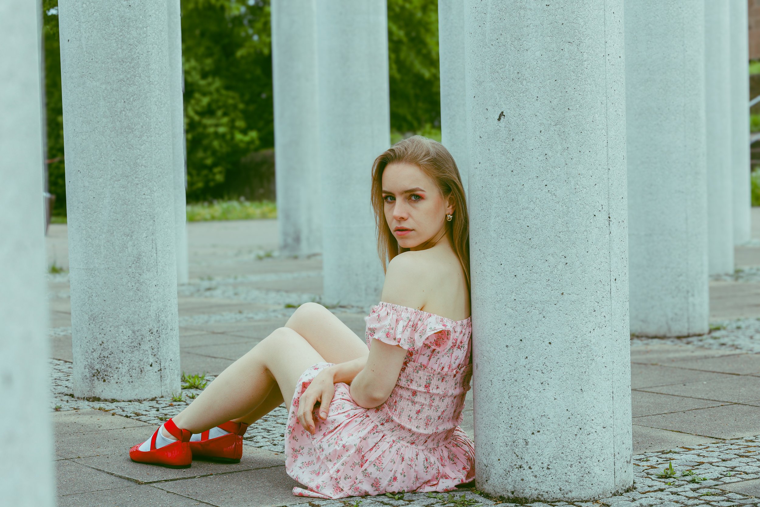 Eine junge Frau sitzt zwischen mehrere weiße, runde Betonsäulen im Freien, trägt ein rosa-blumiges Kleid, rote Schuhe und hat lange braune Haare, blickt nach vorne, umgeben von grünen Bäumen.