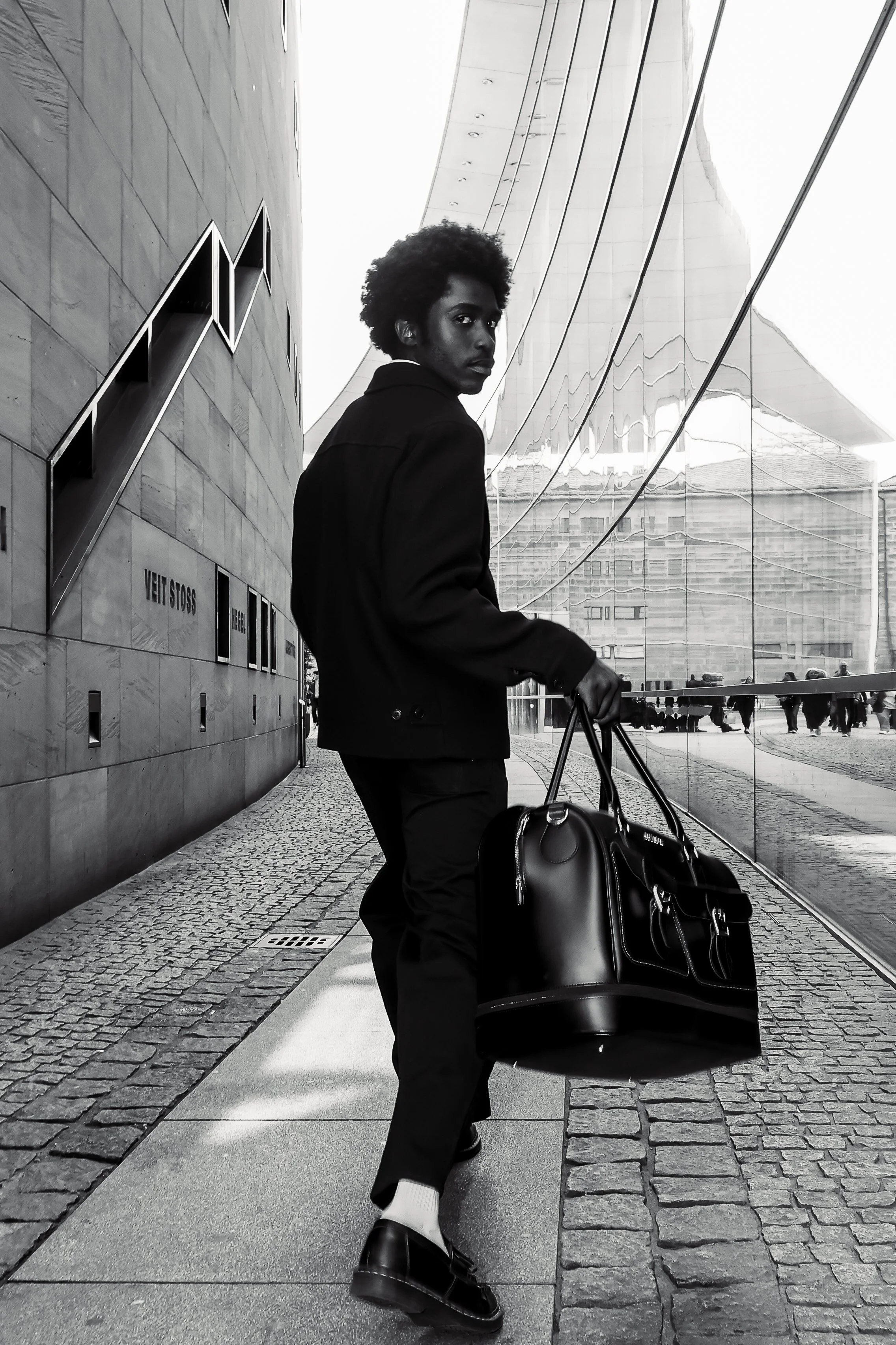 Ein junger Mann mit Afro-Haare steht neben einem schwarzen Koffer vor einem modernen Gebäude mit reflektierender Glasfassade, das auch auf der gegenüberliegenden Seite sichtbar ist, in einer urbanen Umgebung.