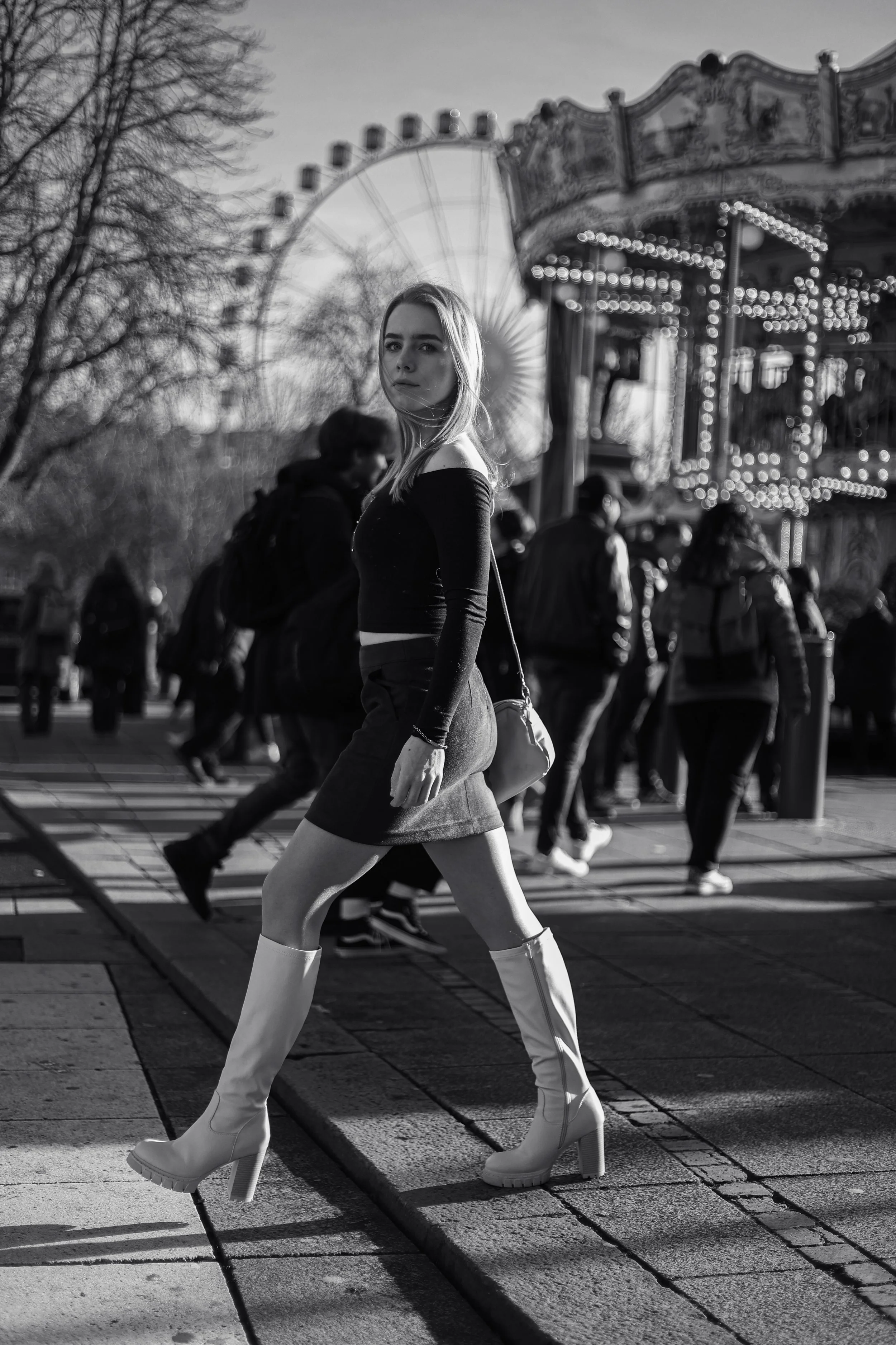 Schwarz-weiß-Foto einer jungen Frau mit langen Beinen, die auf der Straße in Bewegung ist, während im Hintergrund ein Karussell und Menschen unterwegs sind.
