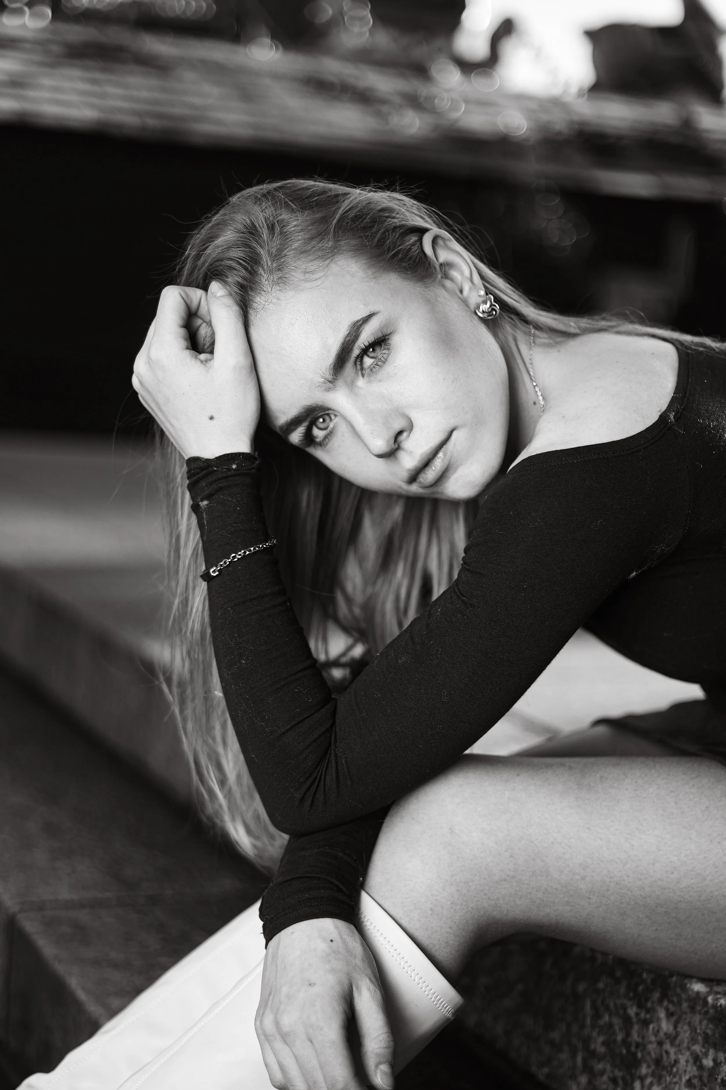 Schwarz-Weiß-Foto einer jungen Frau, die auf einer Oberfläche sitzt und ihre Hand im Haar hält, mit einem nachdenklichen Ausdruck. Sie trägt Ohrringe und ein langärmeliges schwarzes Oberteil.