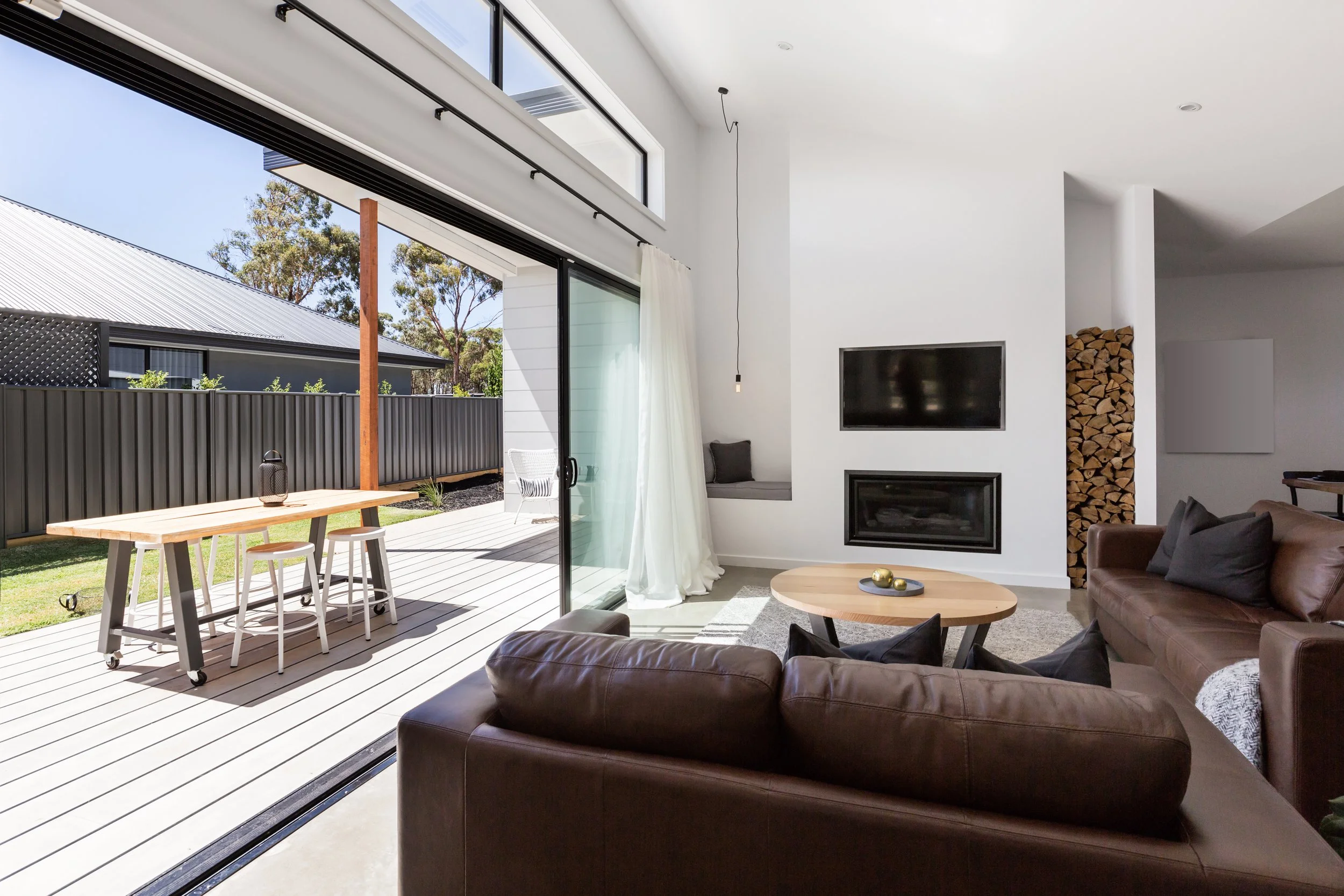 Modern living room with large sliding glass door open to outdoor patio, showing a wooden dining table with stools, white curtains, TV mounted on white wall, stacked firewood, and a brown leather sofa.