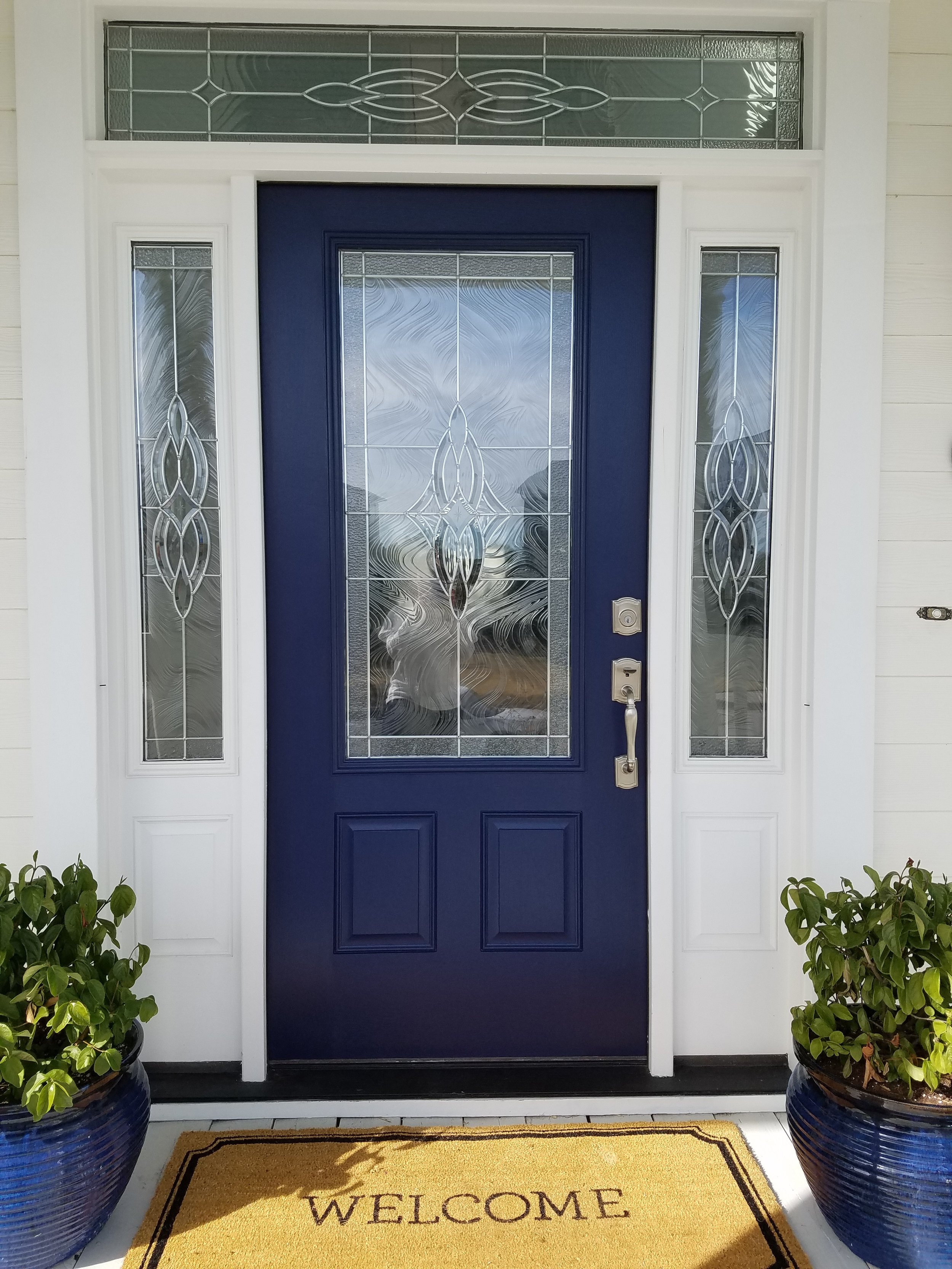Front door with a blue door and decorative glass panels, flanked by glass sidelights and a transom, with plants in pots on either side and a welcome mat on the porch.