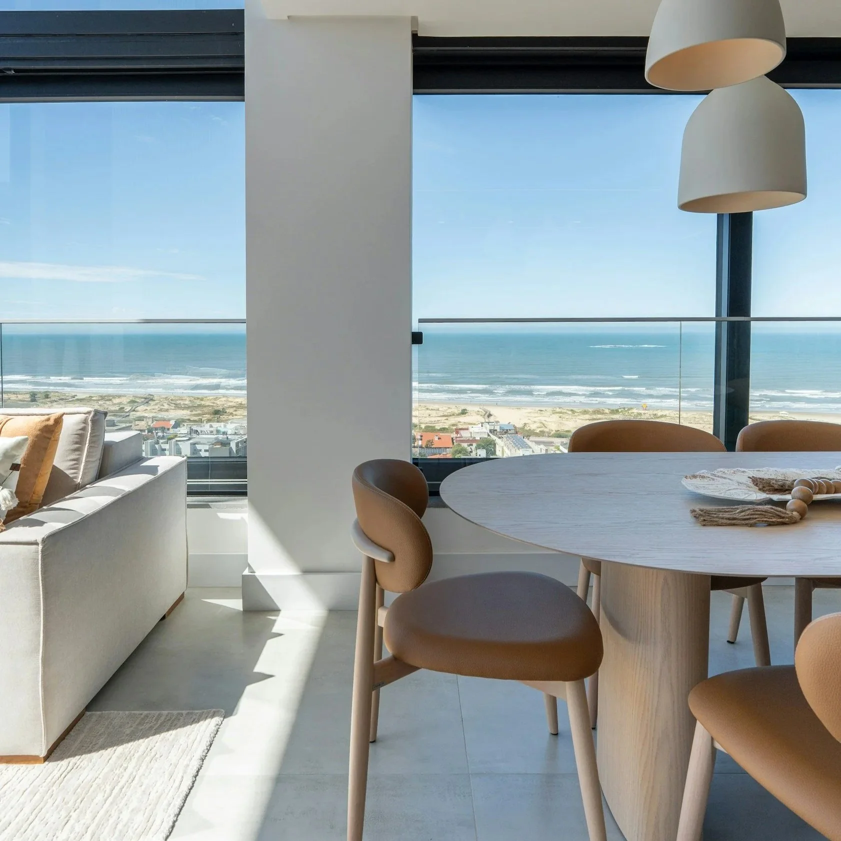 Modern living room or dining area with large floor-to-ceiling windows overlooking a beach and ocean view, a white sofa, round dining table with beige chairs, and pendant lights.