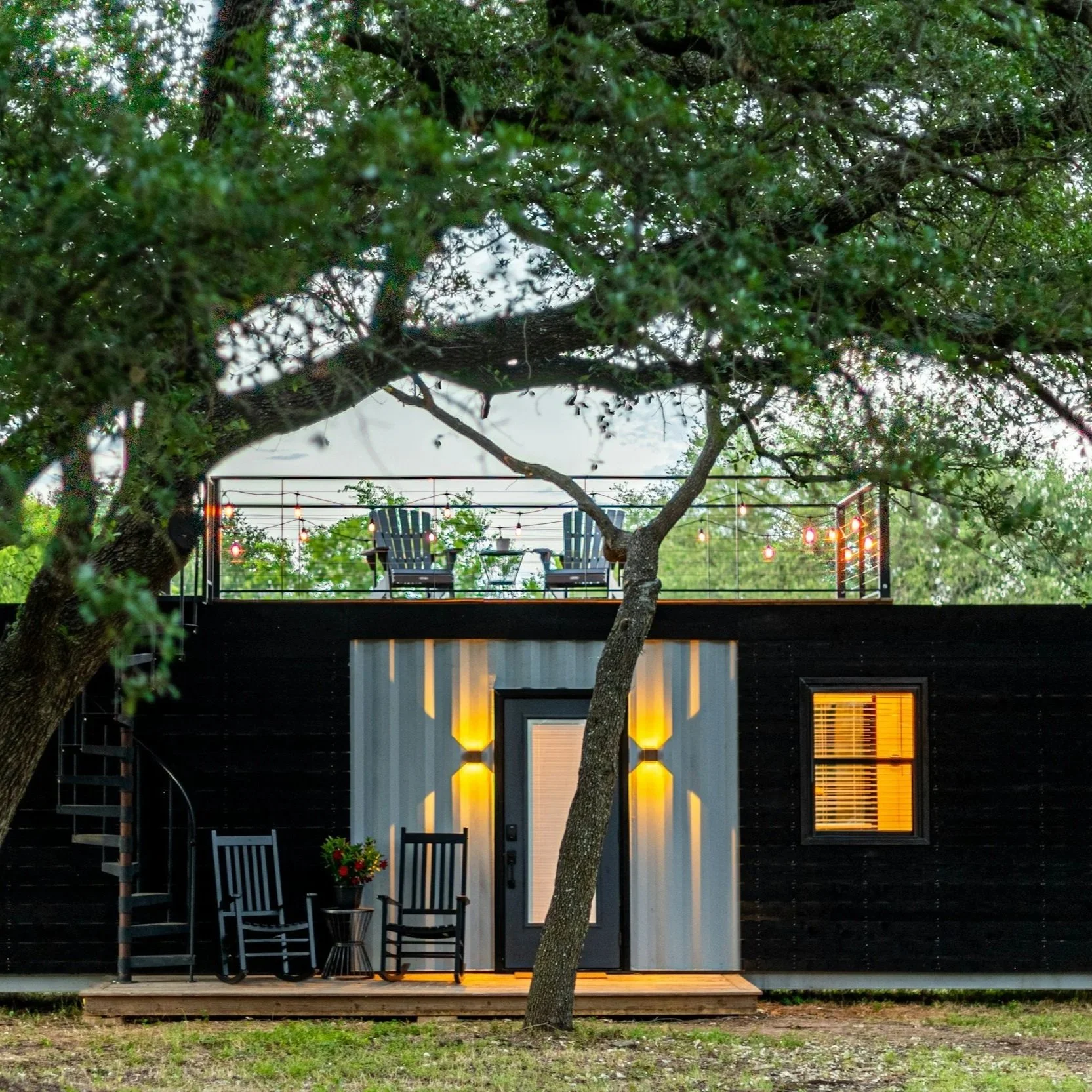 A modern house with a small porch, illuminated by yellow interior lights, is surrounded by trees and outdoor furniture, including chairs, a table, and string lights