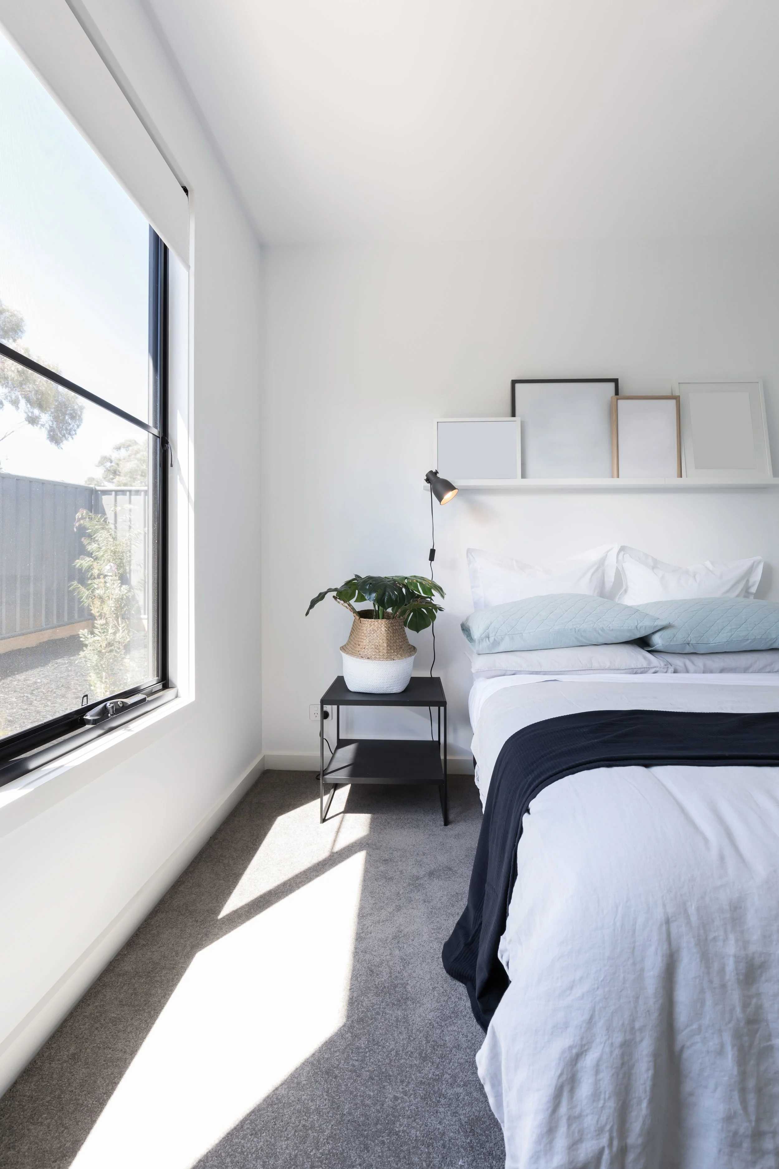 A bedroom with white walls, large window on the left, bed with white and black bedding, a side table with a lampshade, a potted plant, and empty picture frames on a shelf above the bed.