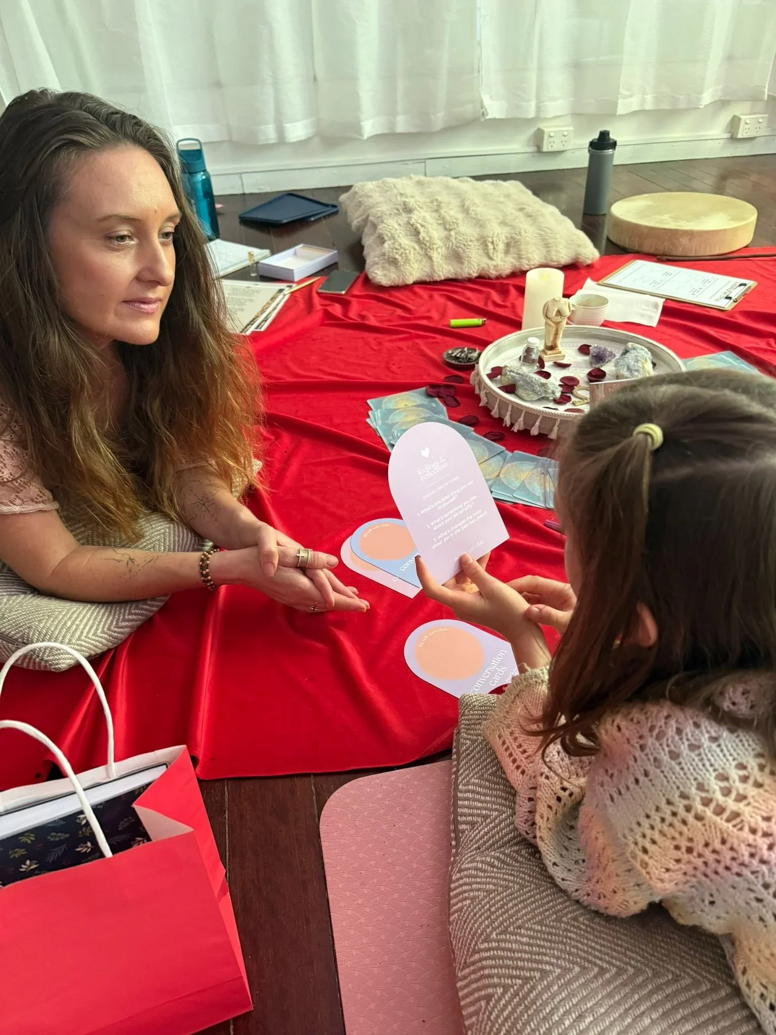 A woman with long wavy hair sitting at a table covered with a red cloth, talking to a young girl with a ponytail. The table has various items including cards, a candle, a tray with stones and rose petals, water bottles, and a notepad. There is a fluffy pillow and a wooden round object on the floor, and a window with white curtains in the background.